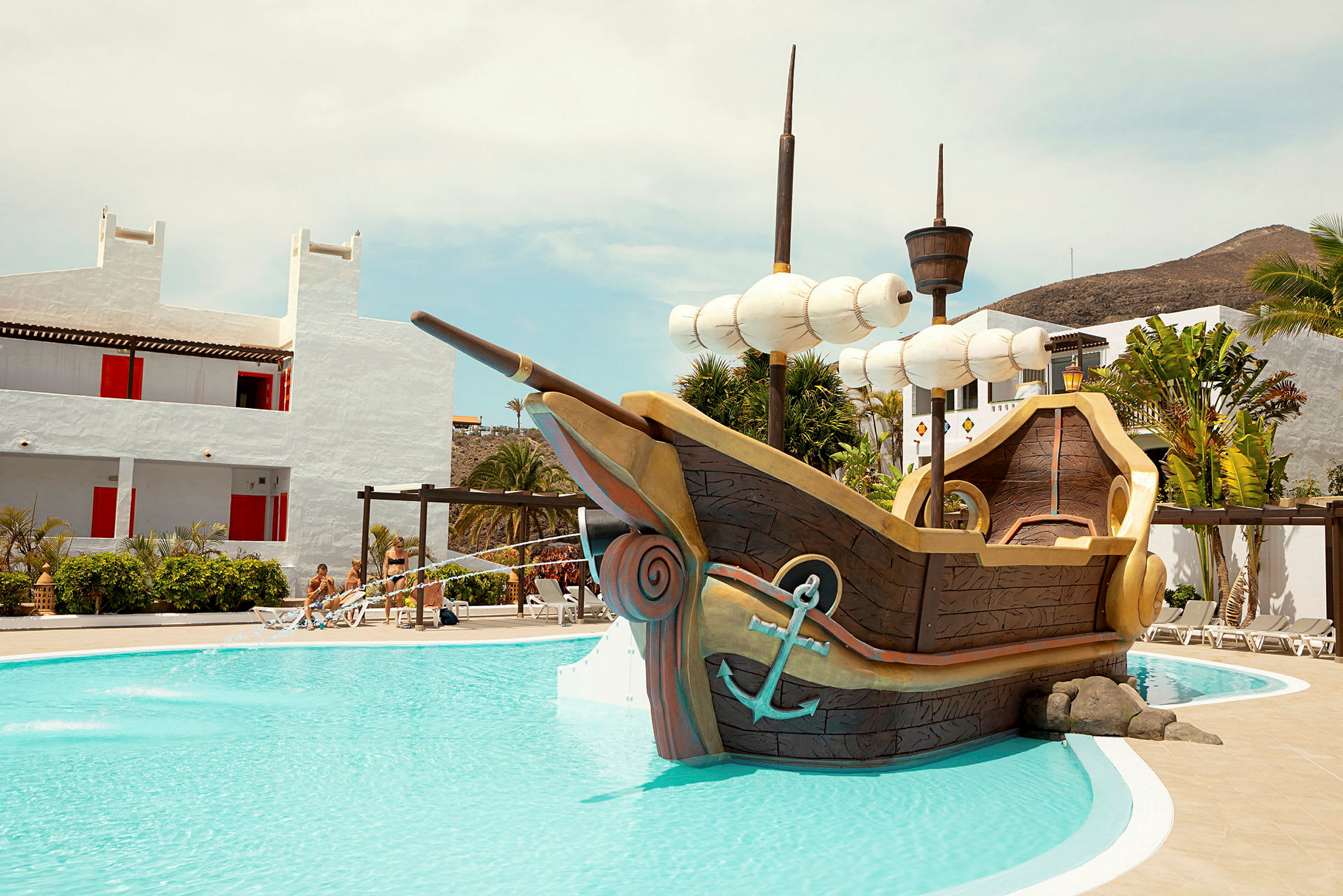 Ett barnvänligt poolområde med ett piratskepp i mitten, omgiven av vitkalkade byggnader. (AI-genererad text)