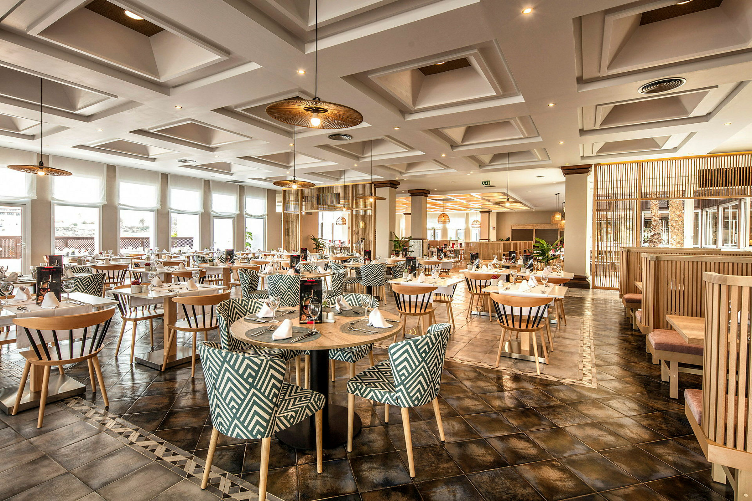 Ett elegant restaurangområde med runda bord och mönstrade stolar, upplyst av taklampor och naturbelyst genom stora fönster. (AI-genererad text)