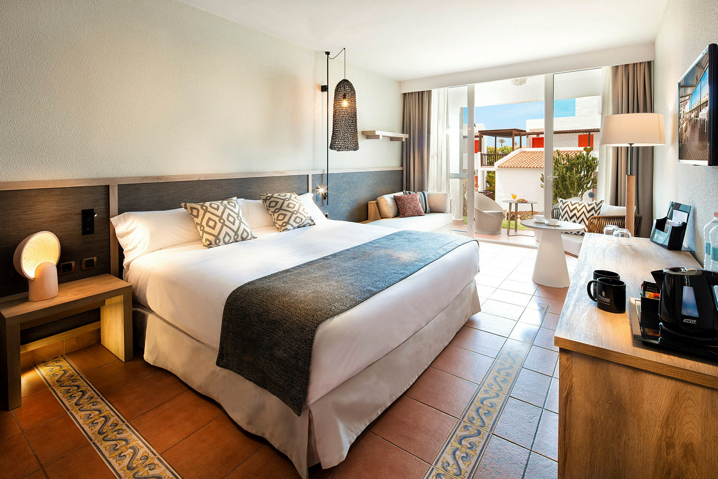 Hotellrum med en stor säng, dekorativa kuddar och modern inredning, öppet mot en ljus balkong med sittgrupp. (AI-genererad text)