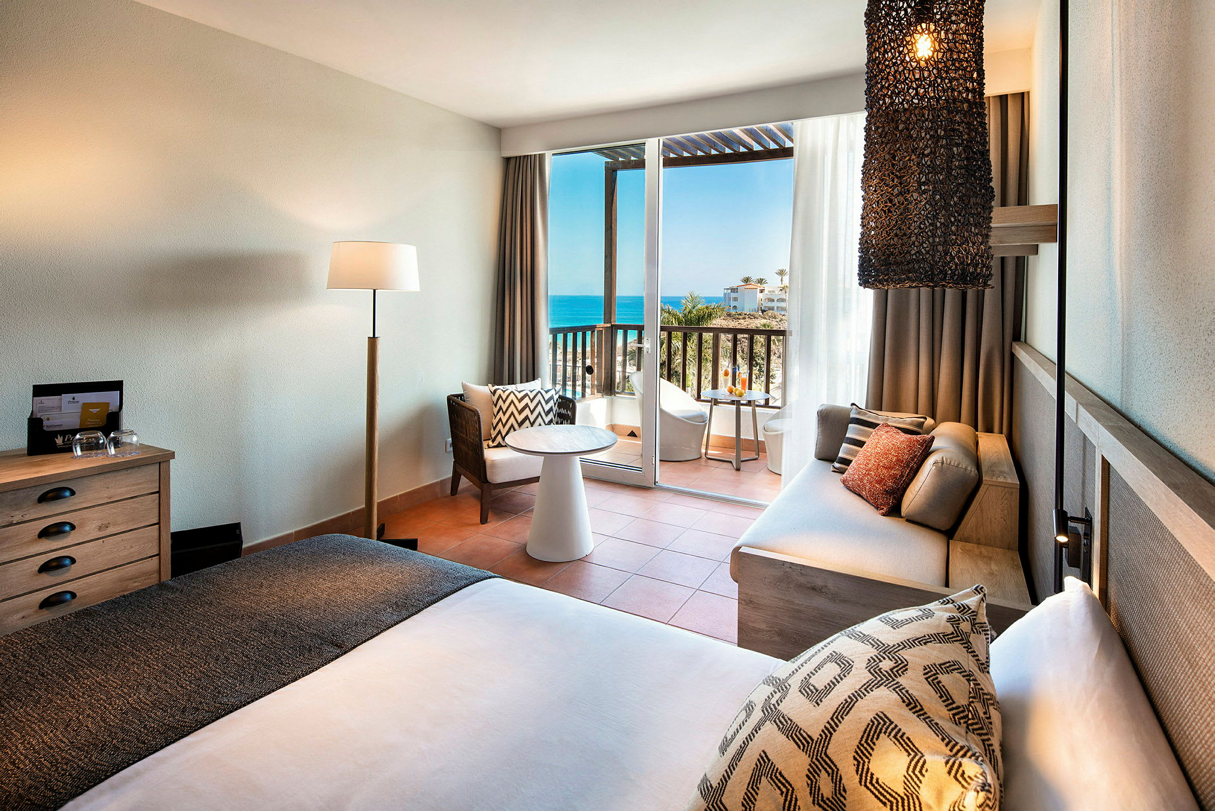 Ett hotellrum med modern inredning som inkluderar en säng, en soffa, ett sidobord, en lampa och en balkong med havsutsikt. (AI-genererad text)