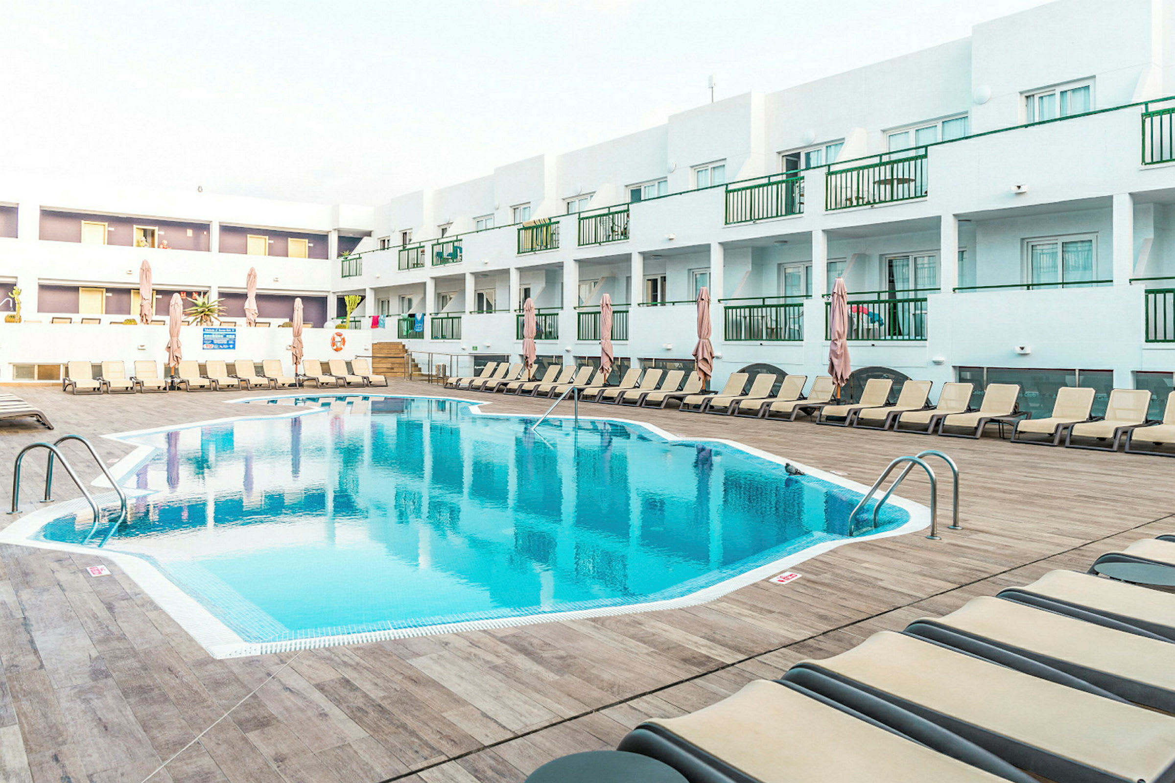 Utomhuspool vid ett hotell, omgiven av solstolar och parasoller, med våningssängar och vita byggnader i bakgrunden. (AI-genererad text)