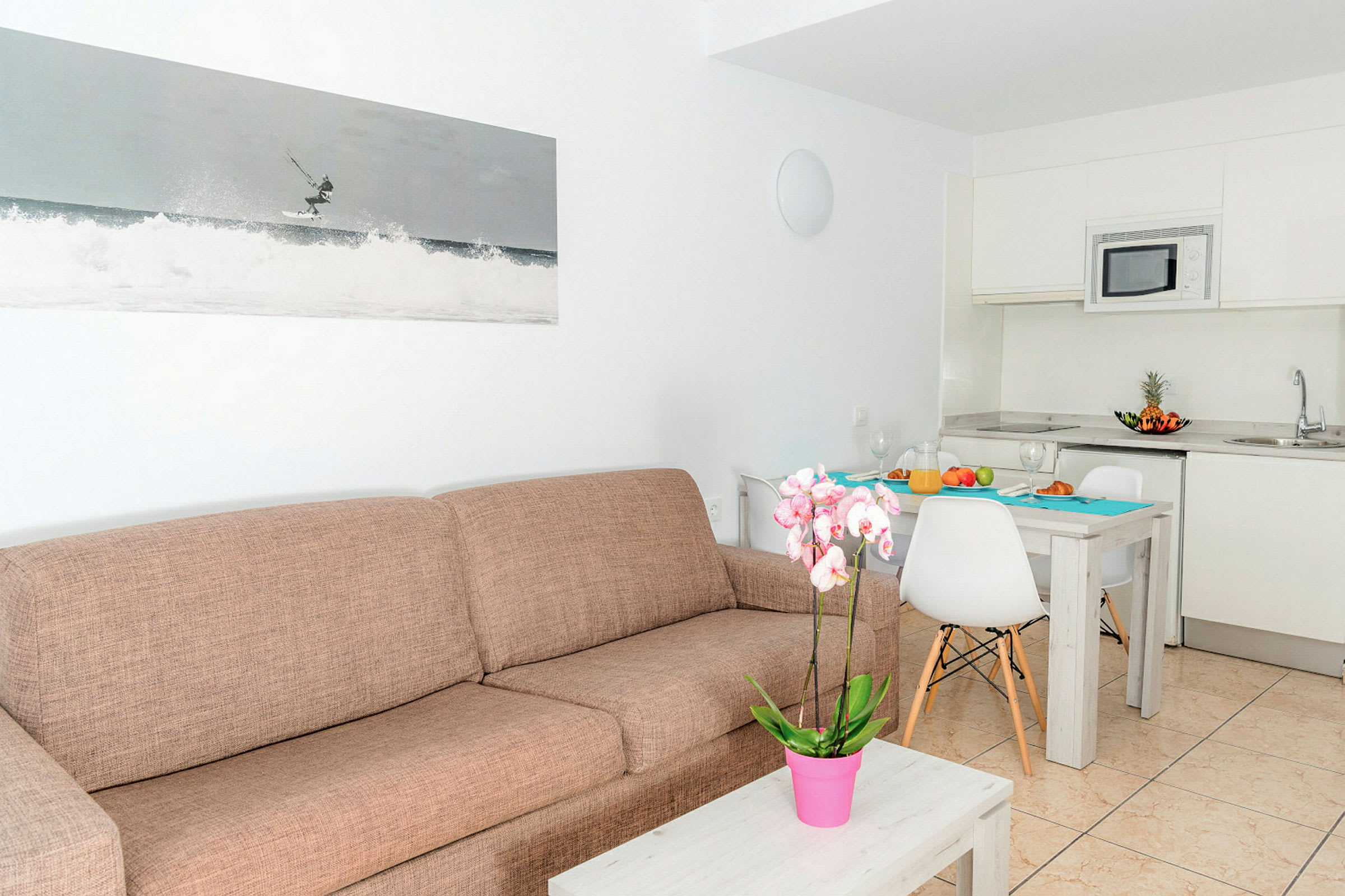 Ett modernt rum med en brun soffa, ett soffbord med en rosa krukväxt, ett kök med vita skåp och en matplats med ett dukat bord. (AI-genererad text)