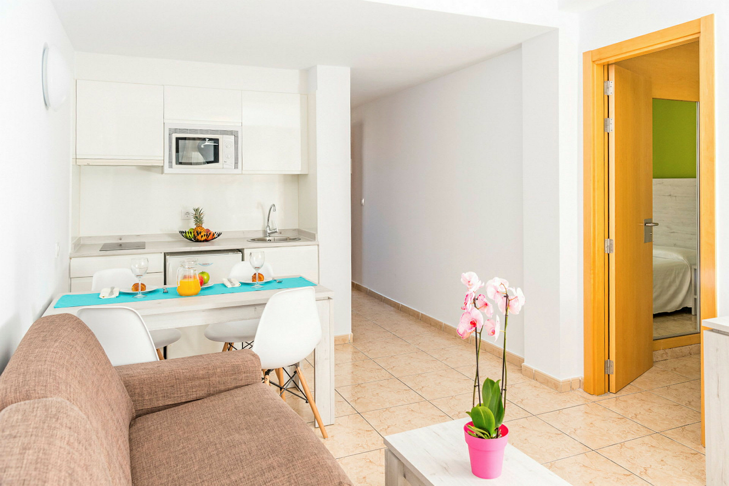Ett modernt kök med matbord och stolar, en brun soffa och en rosa blomkruka på ett soffbord. Ingång till sovrum syns till höger. (AI-genererad text)