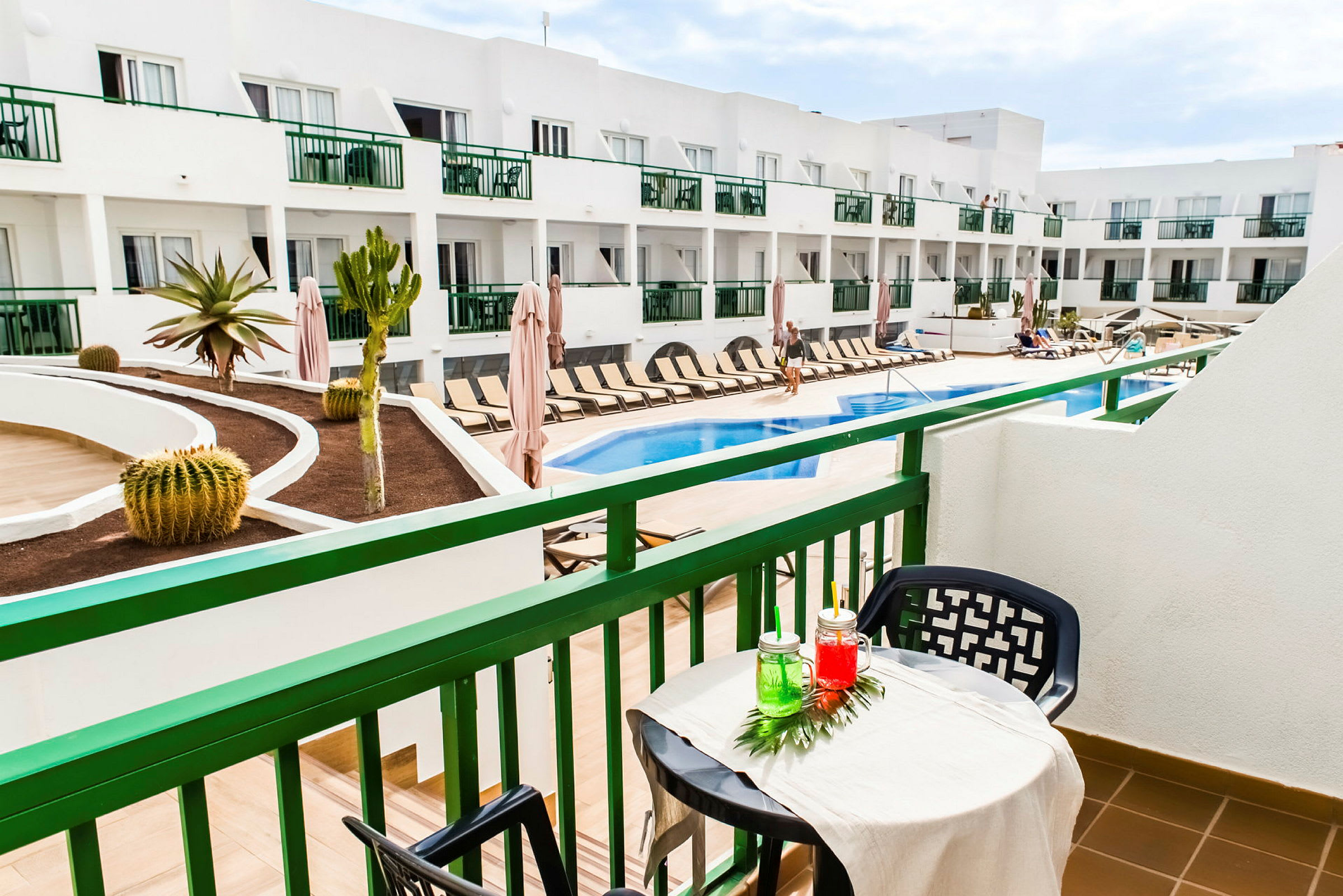 Utsikt från en balkong mot en pool och solstolar på ett hotellområde. (AI-genererad text)