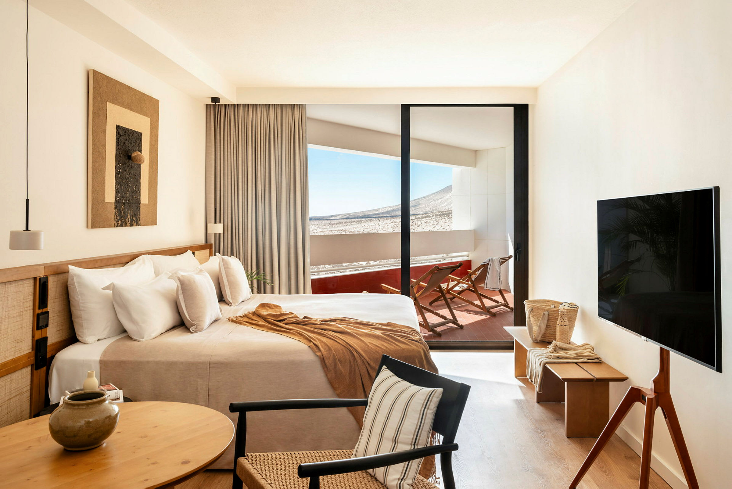 Ett hotellrum med en stor säng, sittplats, TV och utsikt mot en terrass med solstolar och natur i bakgrunden. (AI-genererad text)