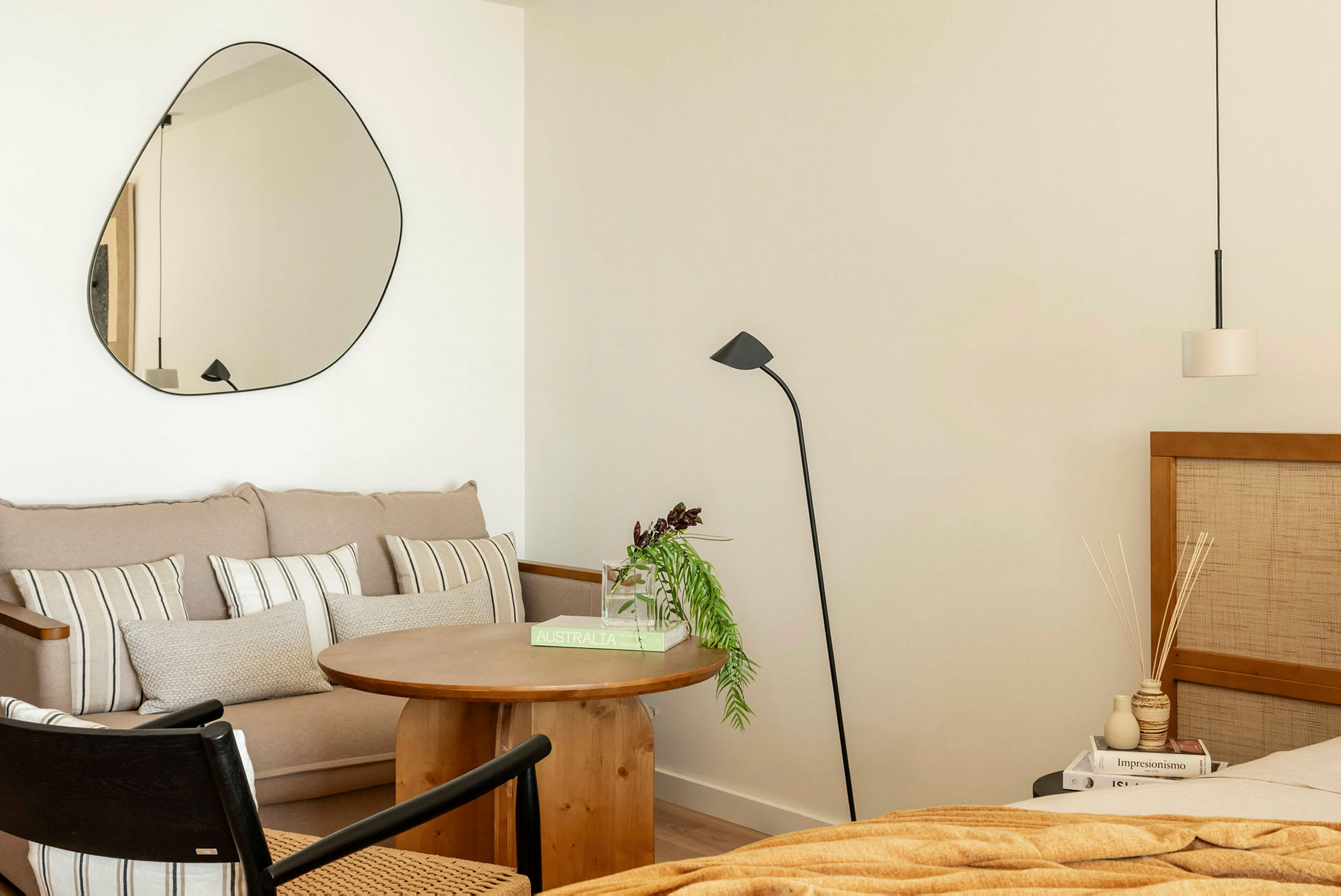 Ett rum med modern inredning. En soffa med kuddar, ett runt träbord, en golvlampa och en spegel på väggen. Till höger syns en säng och ett sängbord. (AI-genererad text)