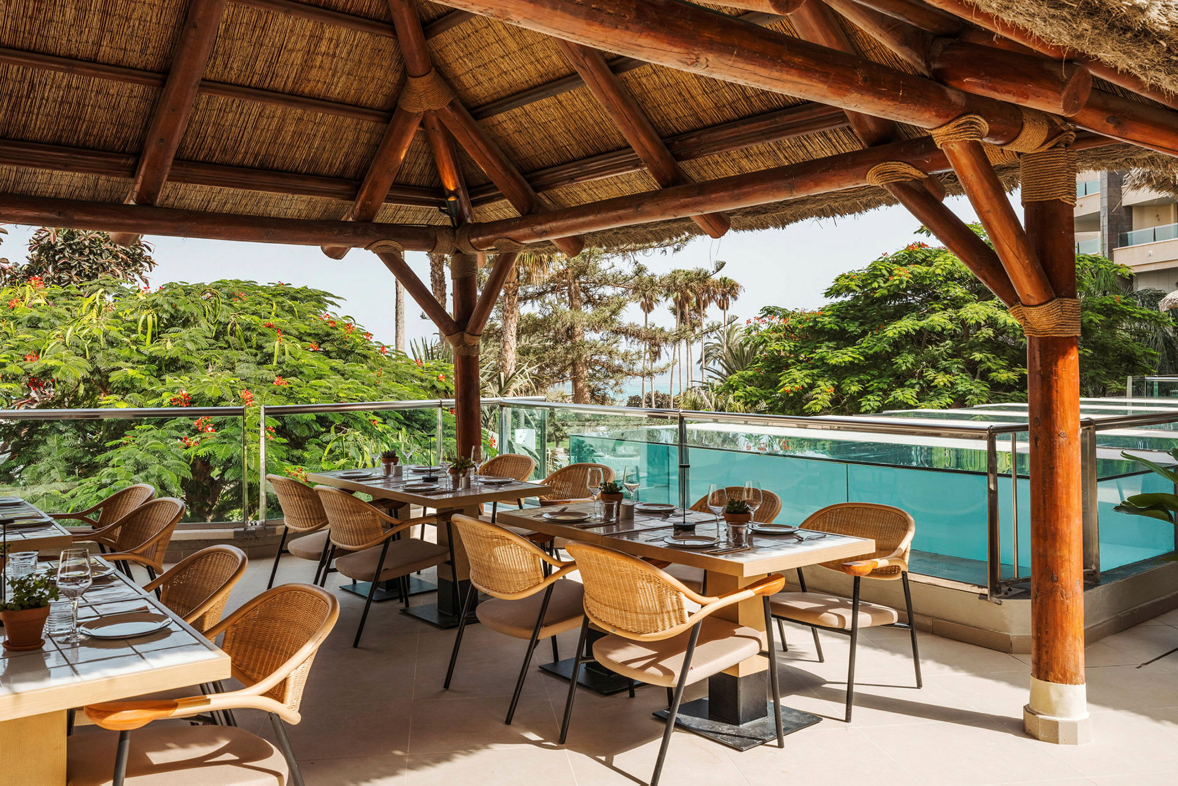 Restaurang med bord och stolar under tak i trä med utsikt mot grönska och pool. (AI-genererad text)