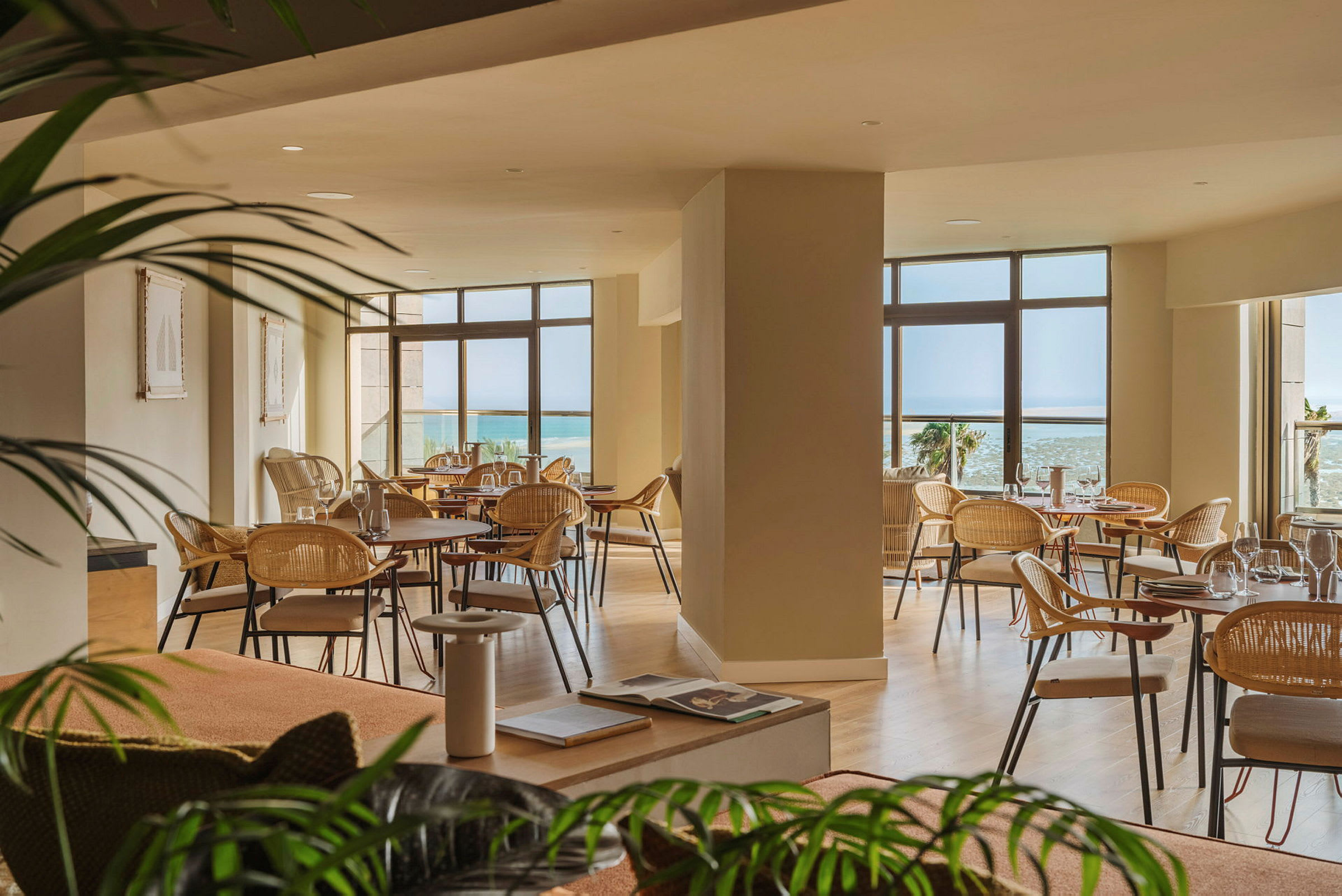 En luftig restaurang med stora fönster som ger utsikt mot havet. Det finns flera bord med stolar och inredningen är ljus och elegant. (AI-genererad text)