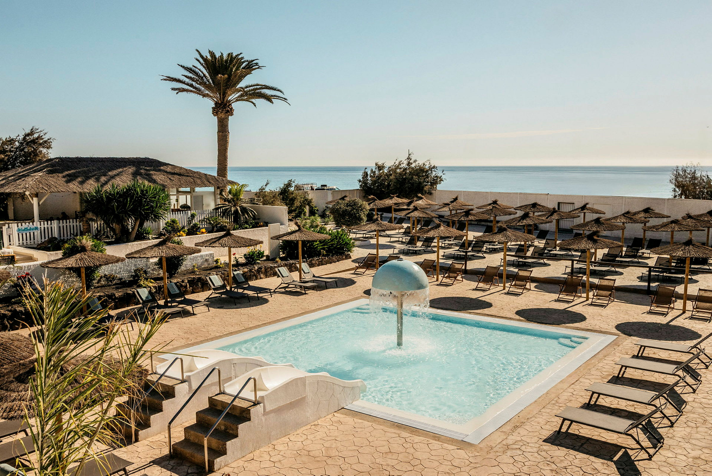 En utomhuspool med en vattensvamp i mitten, omgiven av solstolar och parasoller. I bakgrunden syns en palm, en byggnad med halmtak och havet. (AI-genererad text)