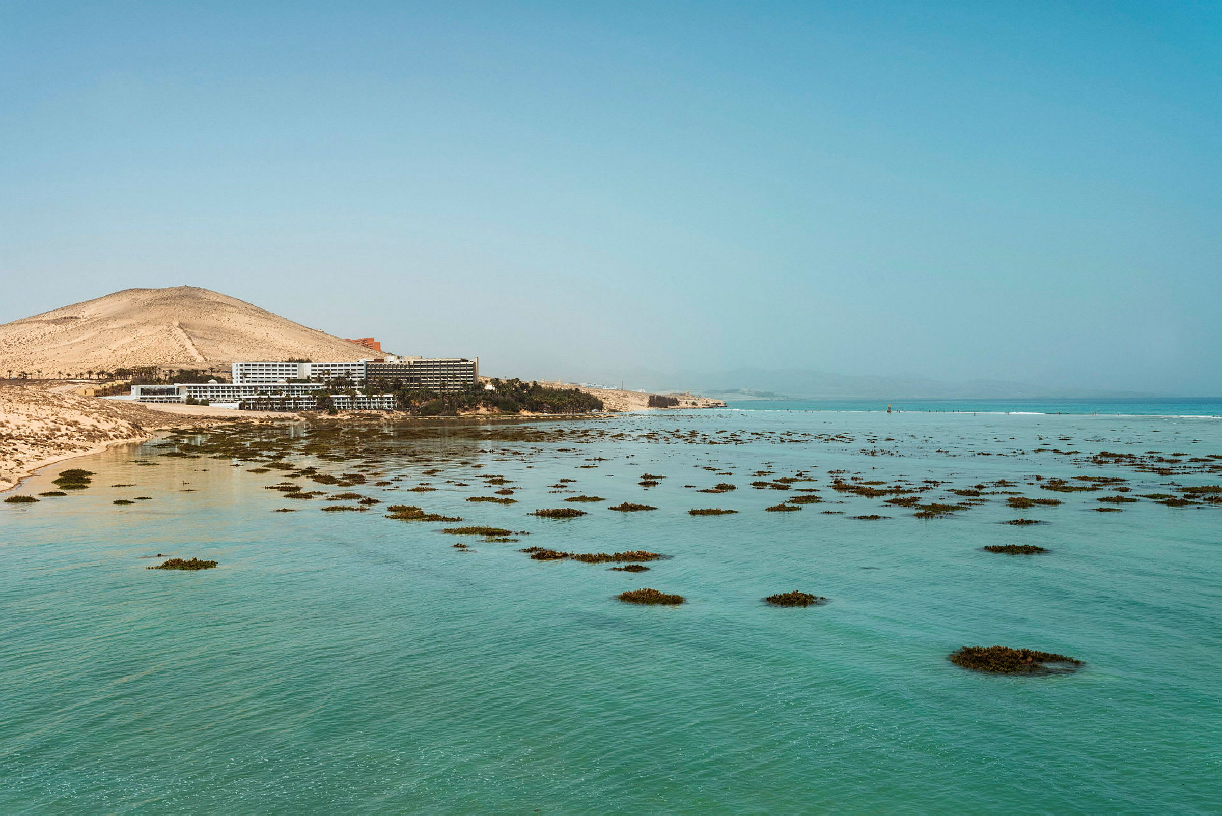 Hotellbyggnader nära en strand med klart blått vatten och spridda kluster av växtlighet. (AI-genererad text)