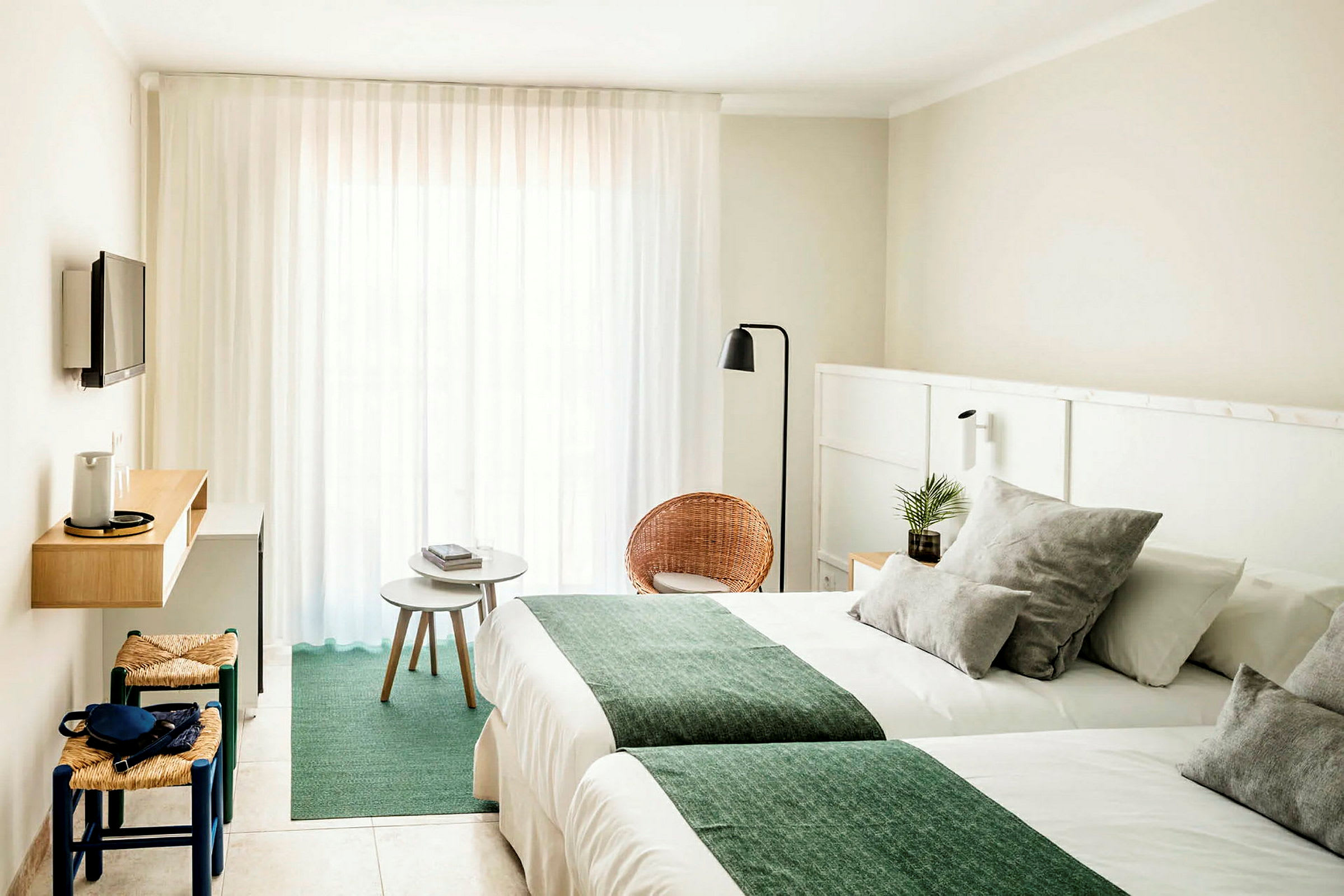 Ett hotellrum med ljusa väggar, två sängar med gröna täcken, en liten sittgrupp med stolar och bord samt en väggmonterad TV. (AI-genererad text)