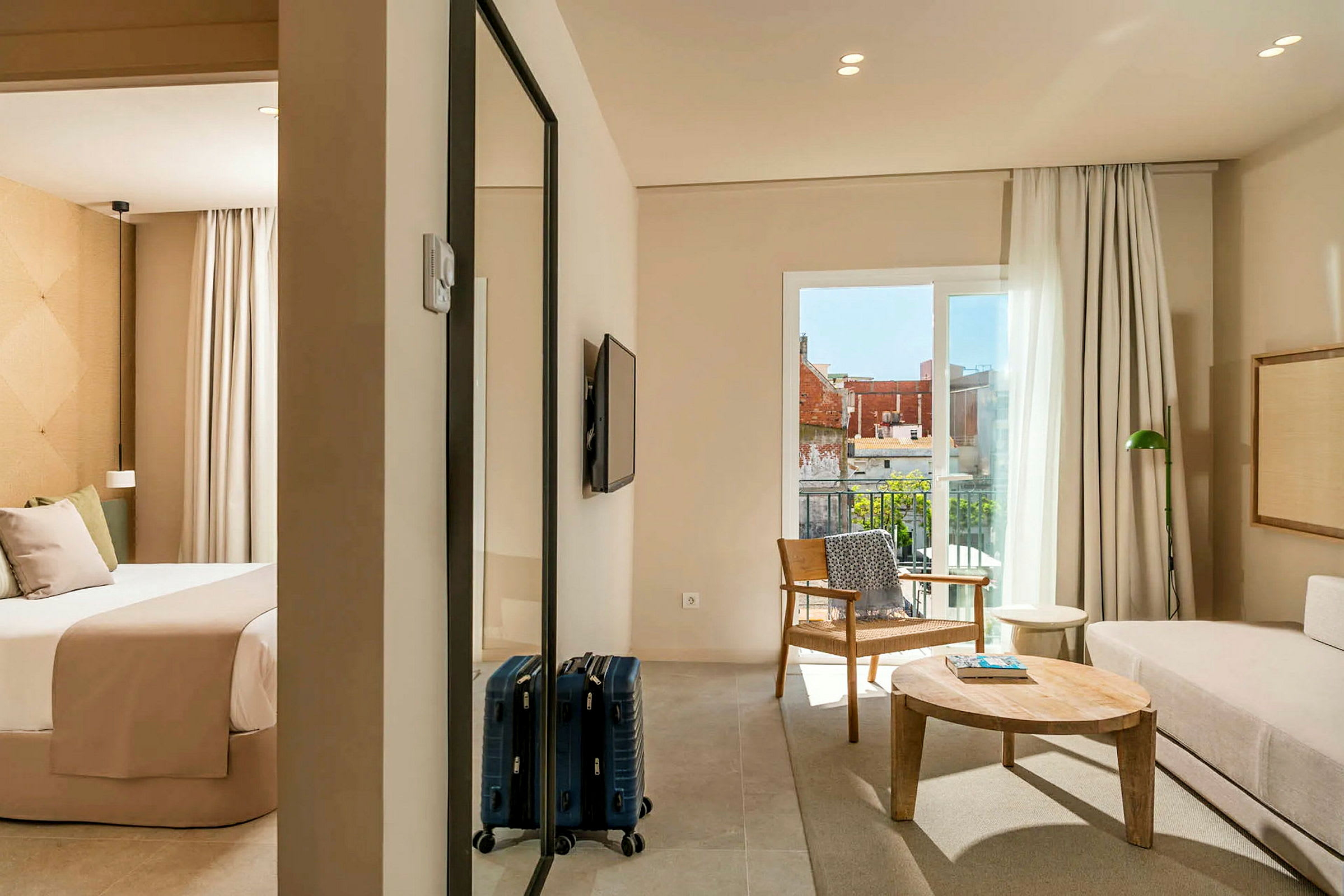 Ett hotellrum med ett sovrum och vardagsrum. Sovrummet har en säng med beige sängkläder, och vardagsrummet har en soffa, ett träbord och en fåtölj. Det finns en balkong med utsikt. (AI-genererad text)
