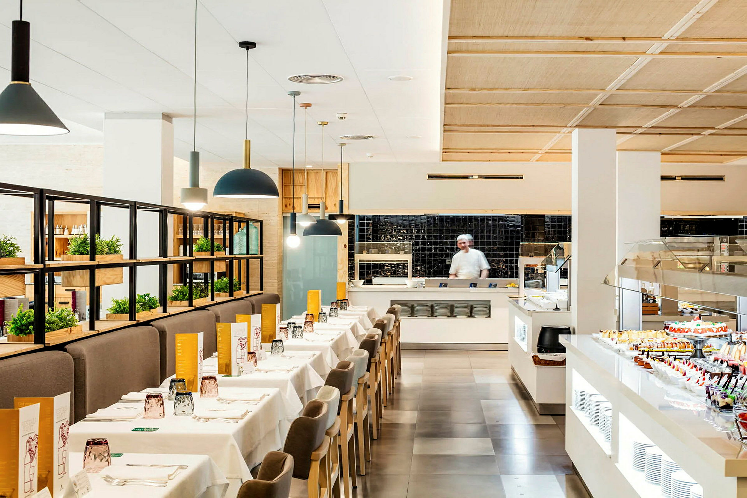 En ljus restaurang med dukade bord, en serveringsbuffé och en kock i köket. (AI-genererad text)