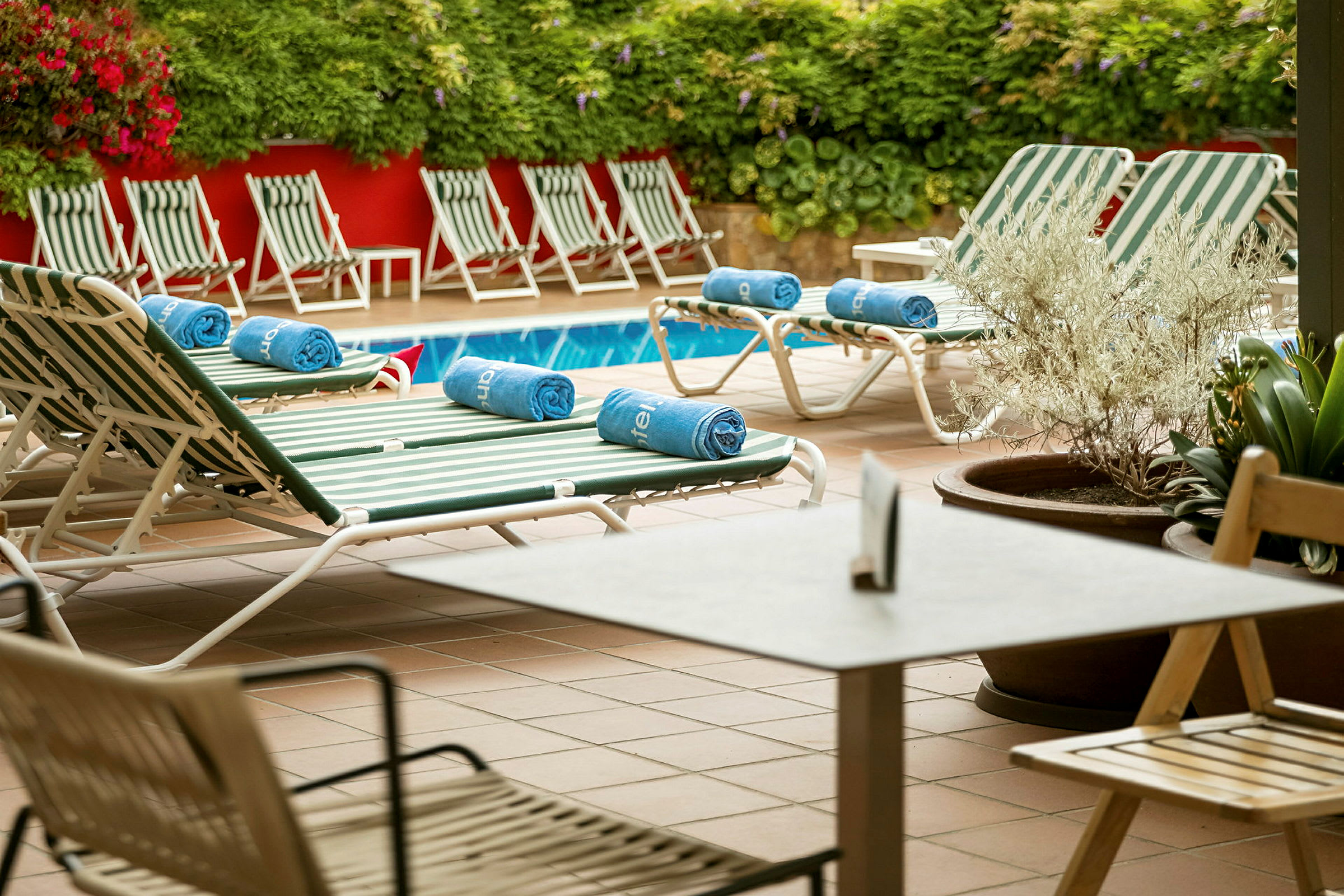 Solstolar och bord vid en pool med grönska och röda väggar i bakgrunden. (AI-genererad text)
