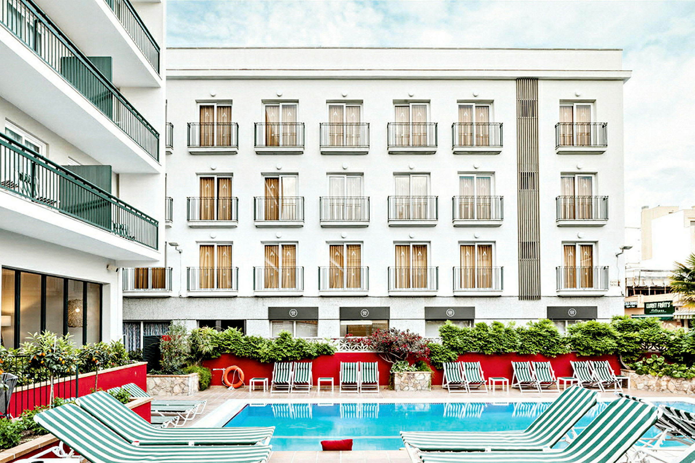 Bild på en hotellbyggnad med vit fasad och flera balkonger. Framför byggnaden finns en utomhuspool med randiga solstolar placerade runt. (AI-genererad text)