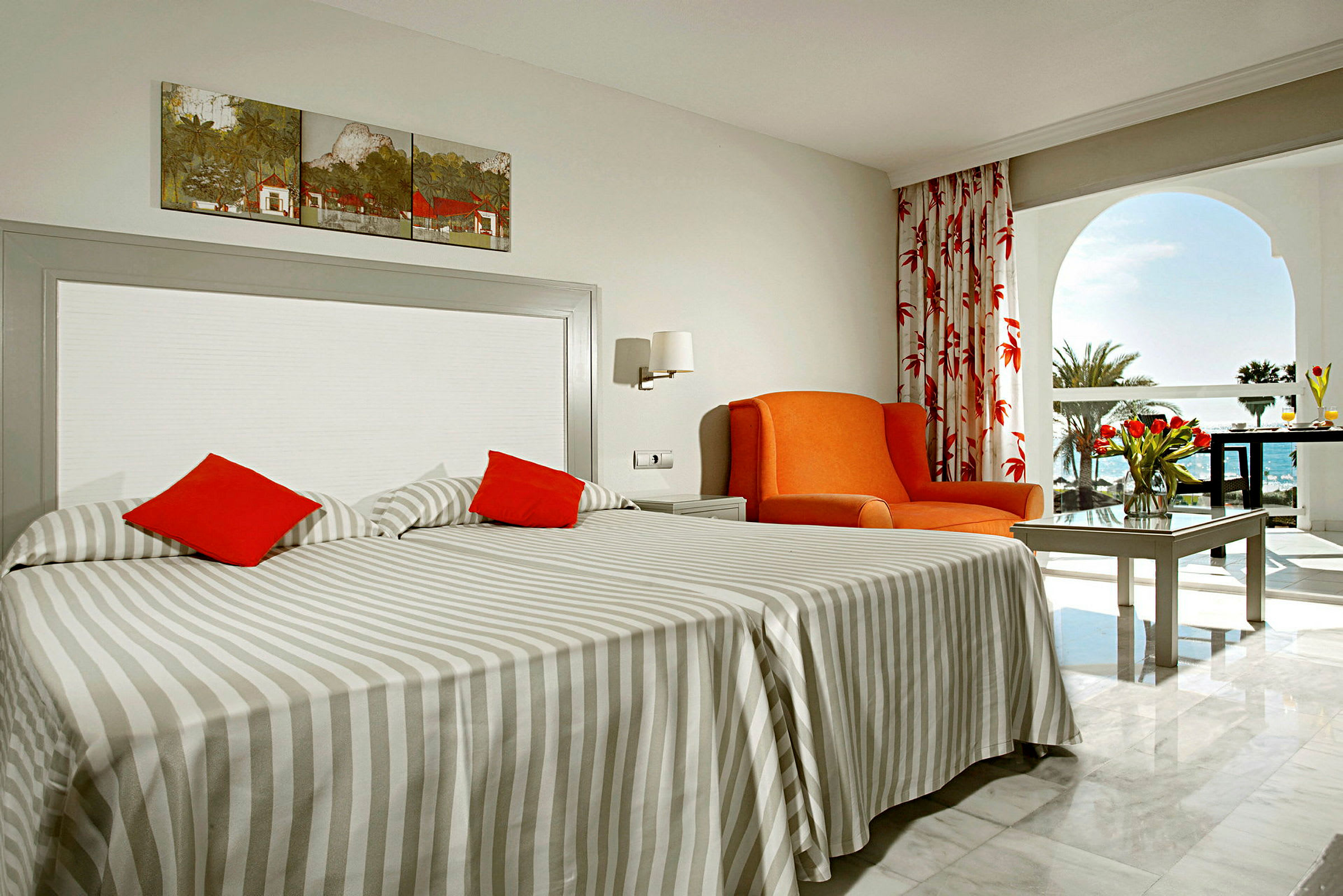 Ett hotellrum med två enkelsängar, en orange fåtölj och ett bord med en bukett tulpaner. Bakom rummet syns en balkong och utsikt över havet. (AI-genererad text)