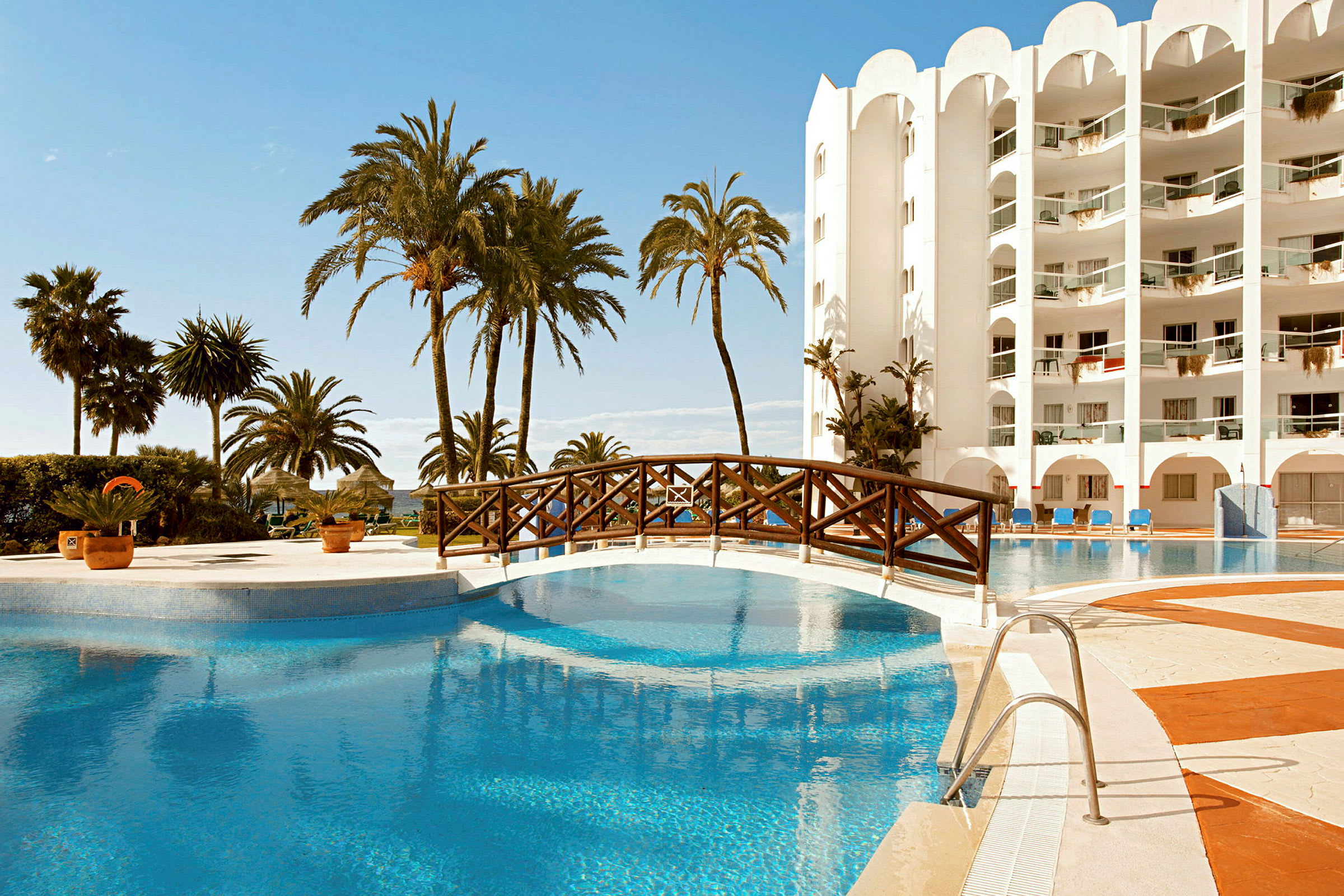En pool vid ett hotell med palmer och en träbro över vattnet. (AI-genererad text)