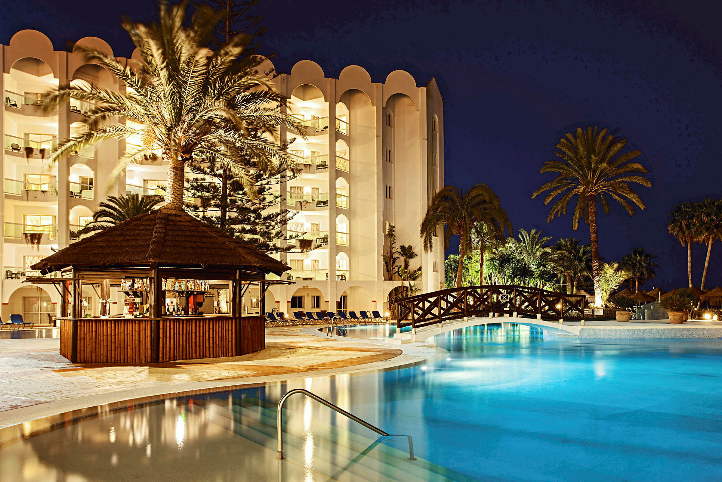 Ett upplyst hotellkomplex vid en pool på kvällen med palmer och en träbro. (AI-genererad text)