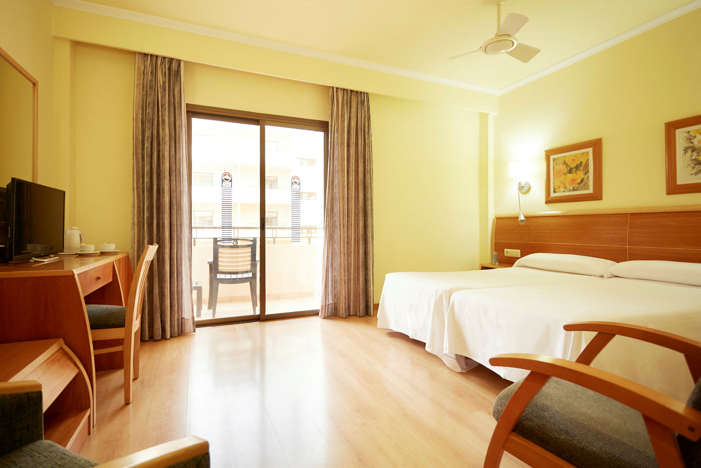 Ett ljust hotellrum med två sängar, skrivbord, stol, och balkong med glasdörrar. (AI-genererad text)