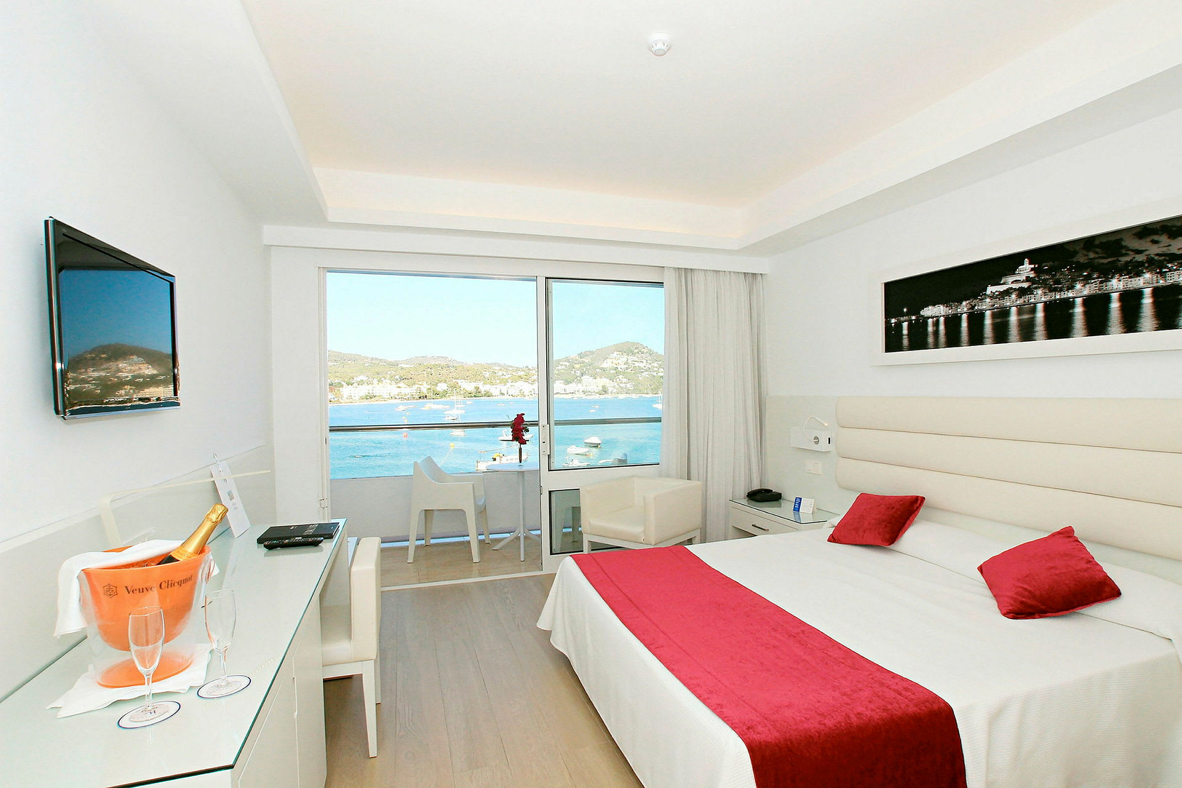 Ett ljust hotellrum med en stor säng, rött överkast och kuddar samt en balkong med utsikt över havet. (AI-genererad text)