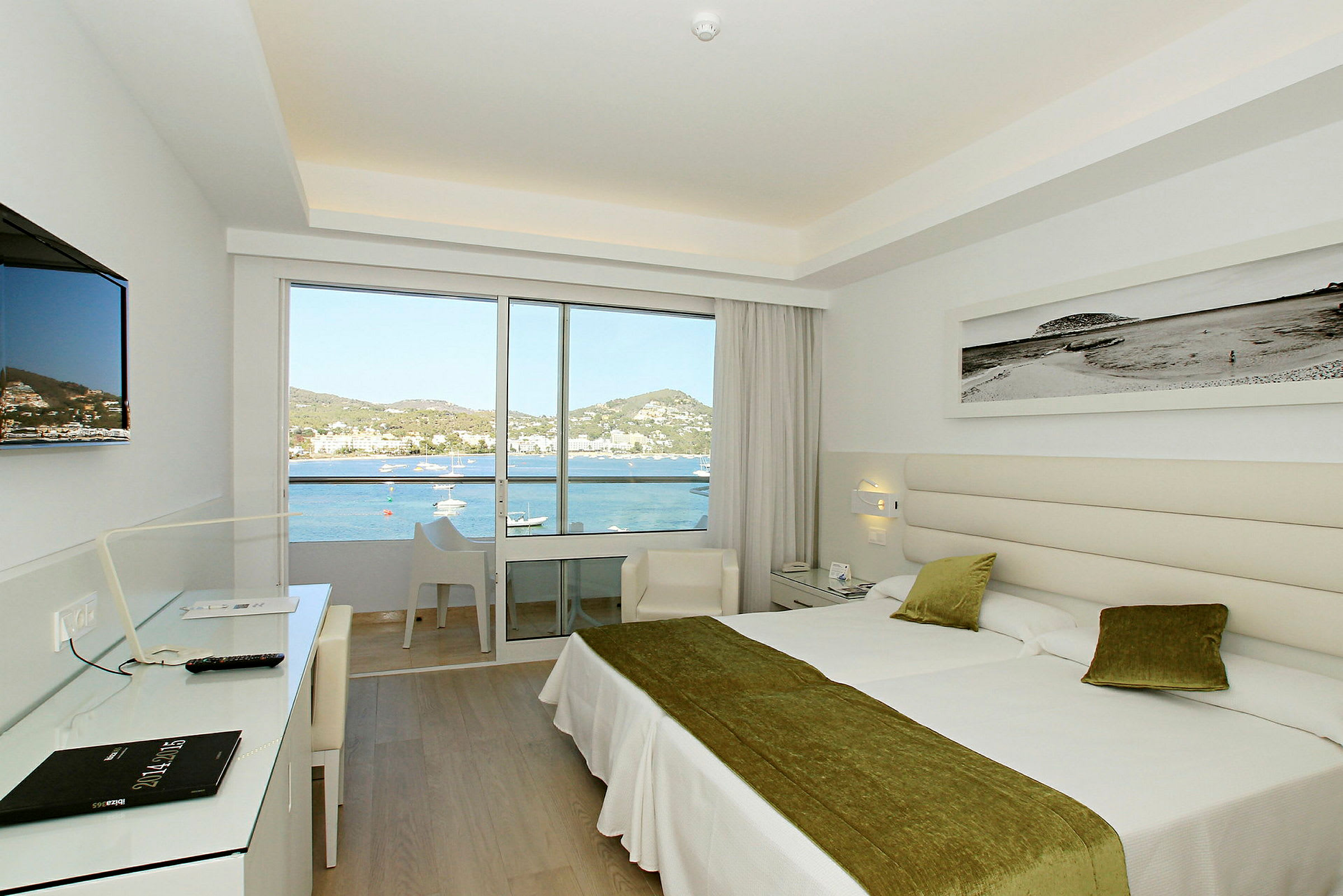 Ett ljus och modernt hotellrum med två sängar och stora glasdörrar som vetter mot en balkong. Utanför syns en vy över havet och kringliggande landskap. (AI-genererad text)