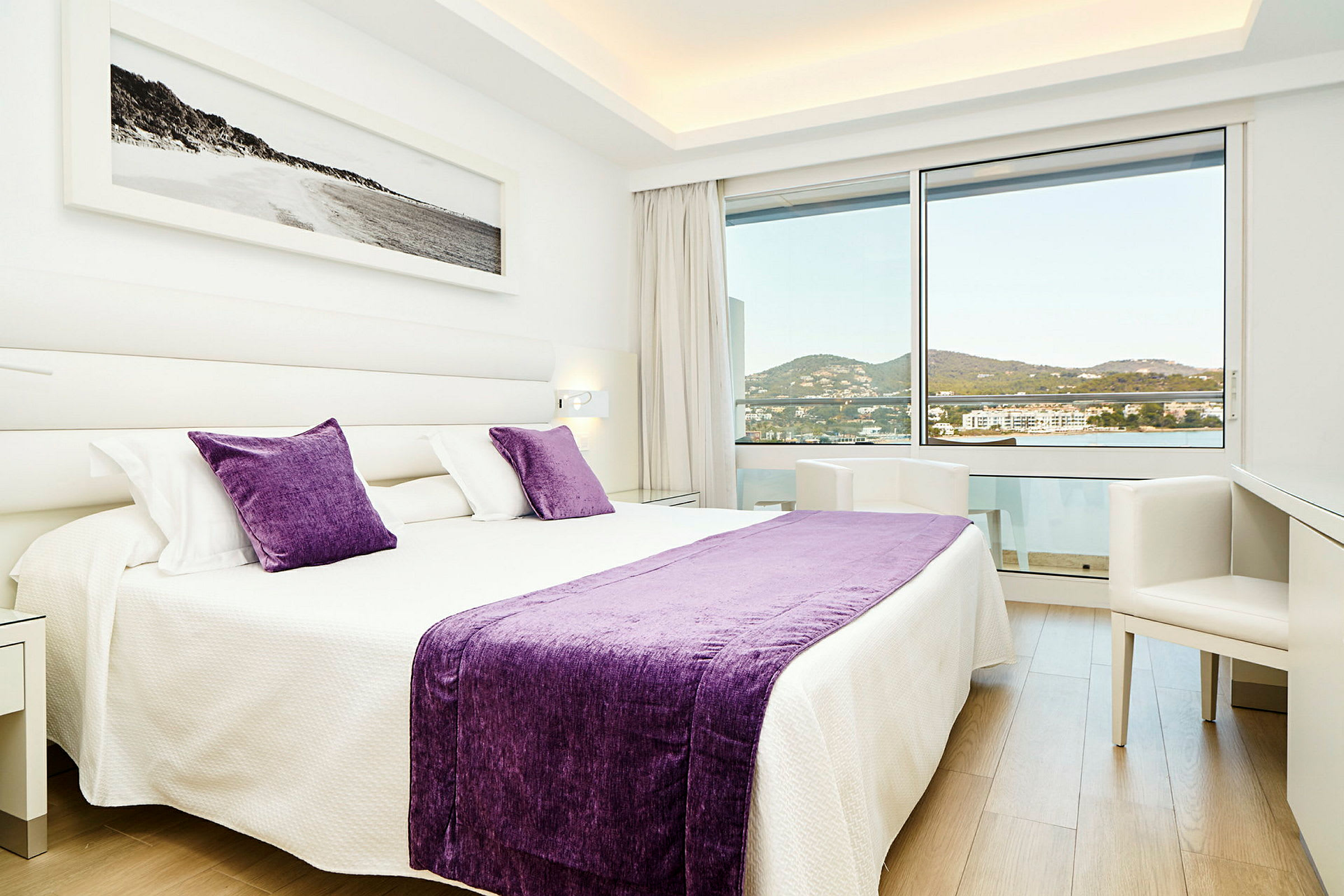 Ett modernt hotellrum med en stor säng dekorerad med lila kuddar och filt, ljusa väggar och ett stort fönster som avslöjar en vacker utsikt över omgivningen. (AI-genererad text)