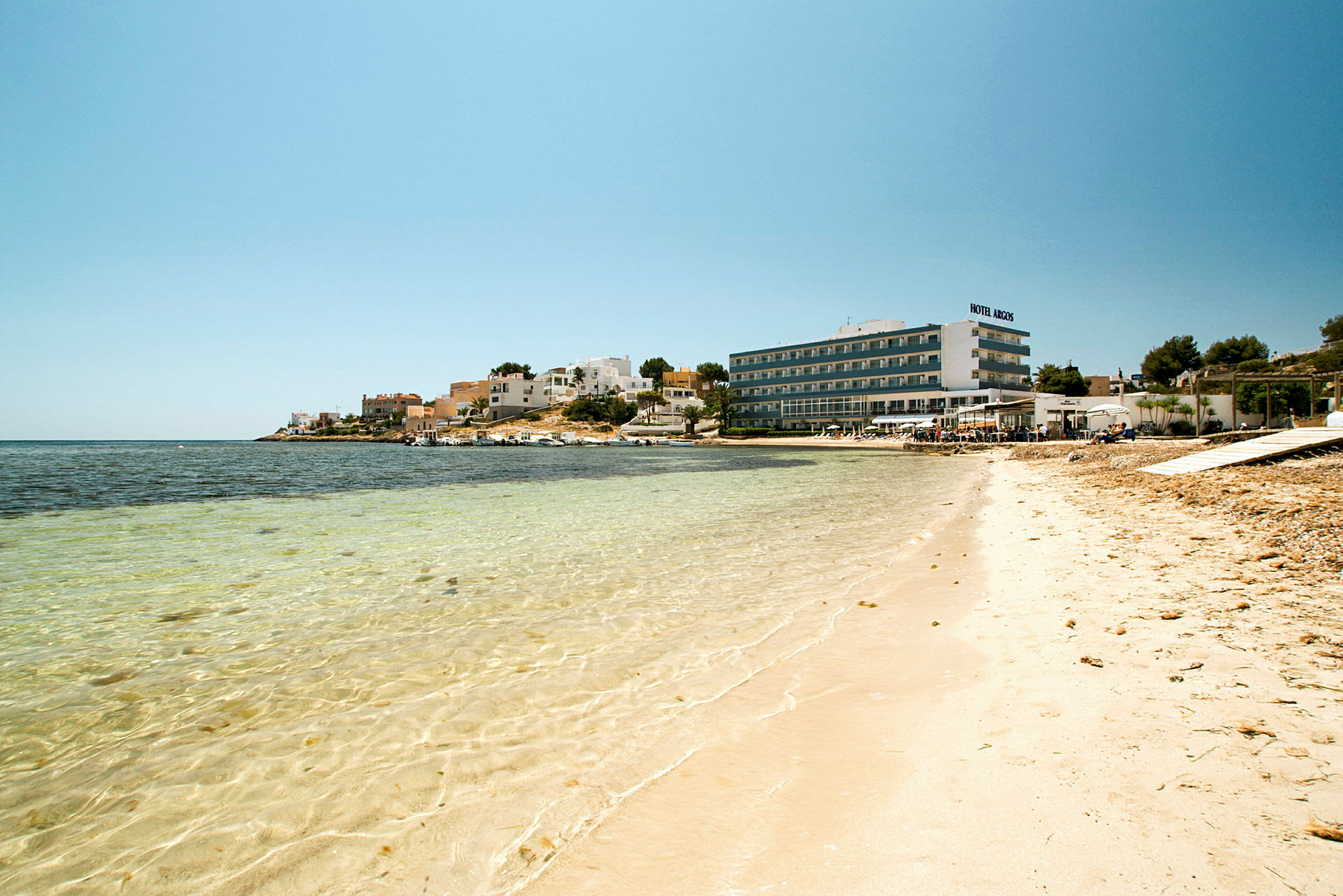 En strand med klart vatten, byggnader och ett hotell i bakgrunden. (AI-genererad text)