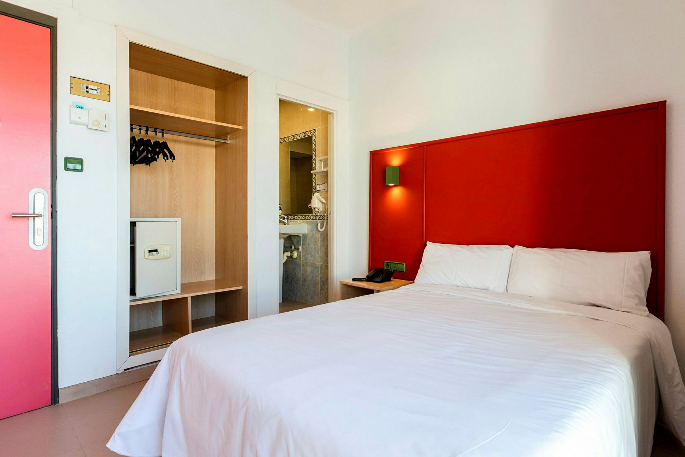 Hotellrum med röd sänggavel, vit säng, och garderob med säkerhetsbox. (AI-genererad text)