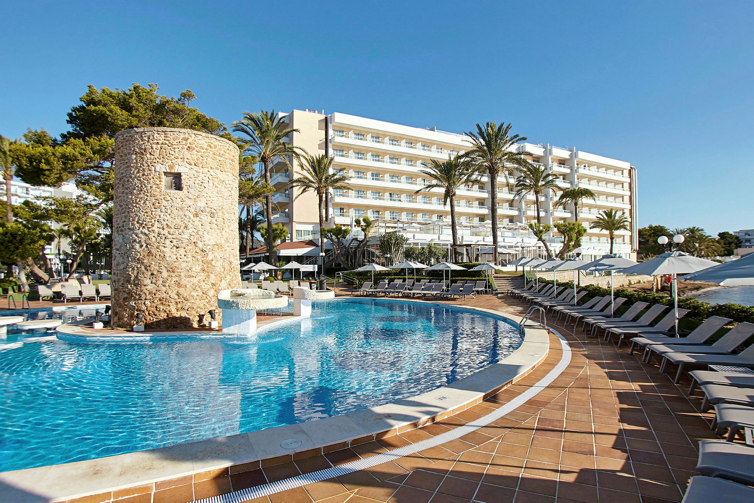 En swimmingpool omgiven av solstolar, med ett stenbyggt torn och ett hotell i bakgrunden. (AI-genererad text)