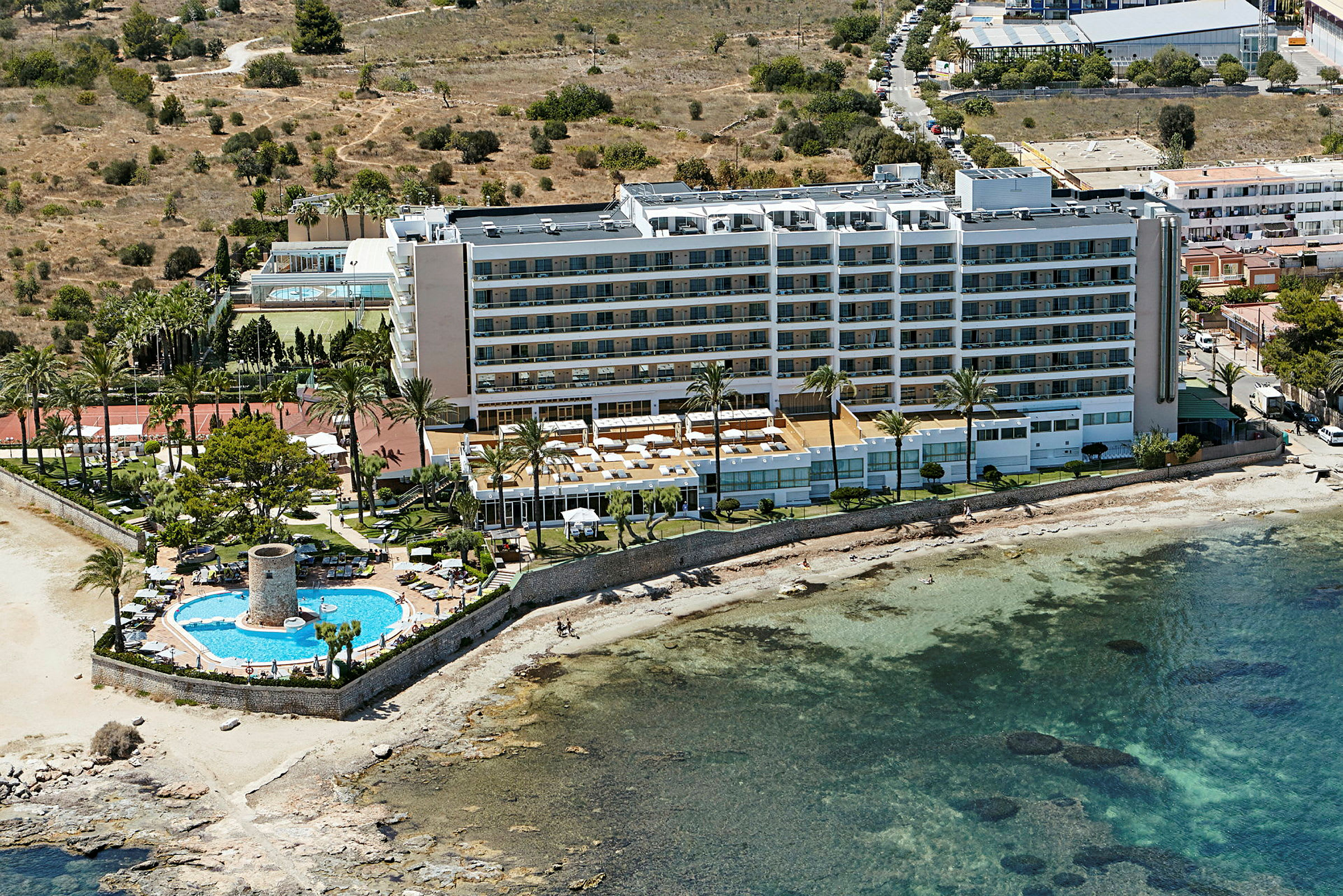 Ett hotell vid havet med en pool och solstolar samt omgivande grönområden. (AI-genererad text)