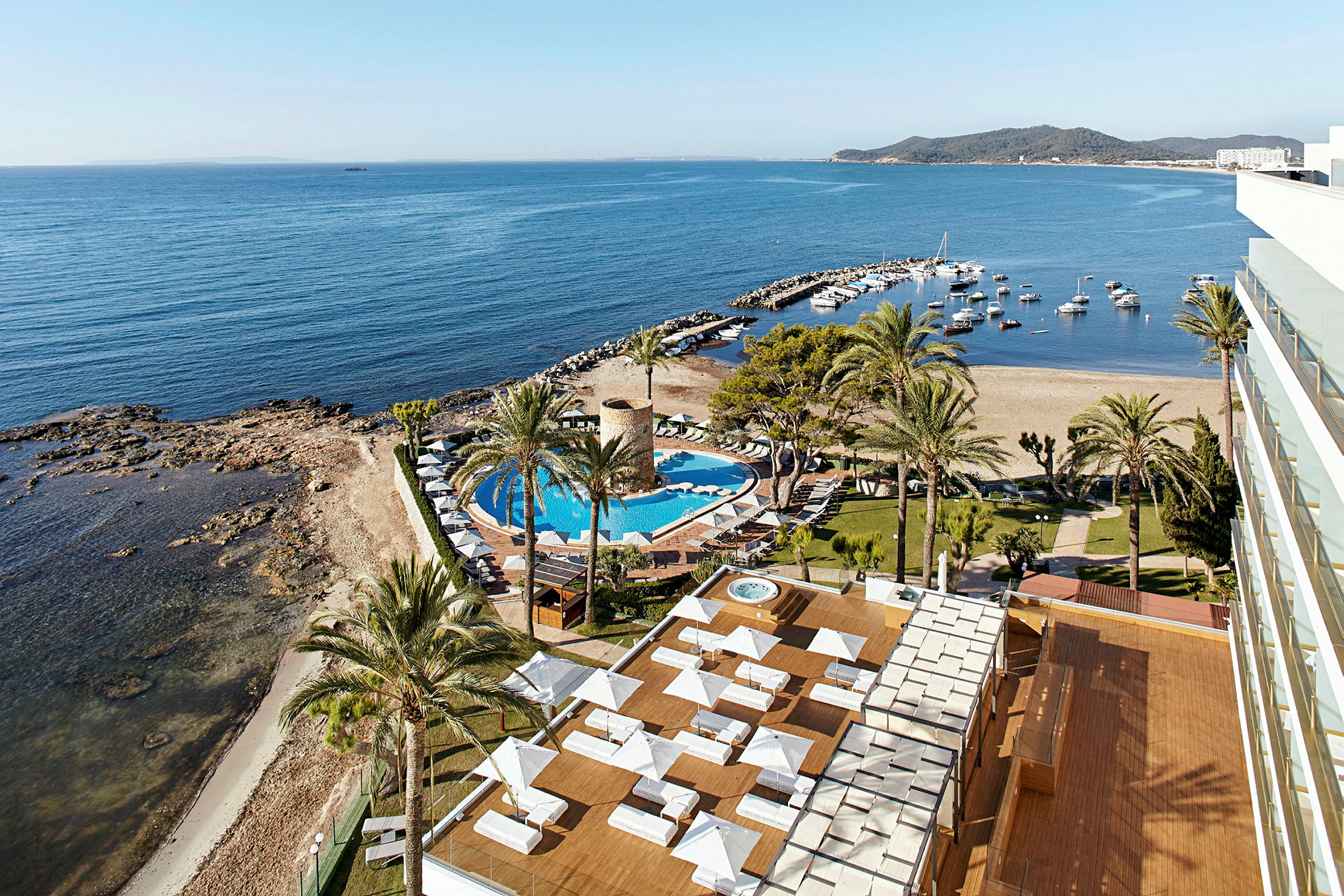 Bildens motiv visar en vy över ett hotellområde vid havet med en pool, solstolar samt en strand och småbåtshamn i bakgrunden. (AI-genererad text)