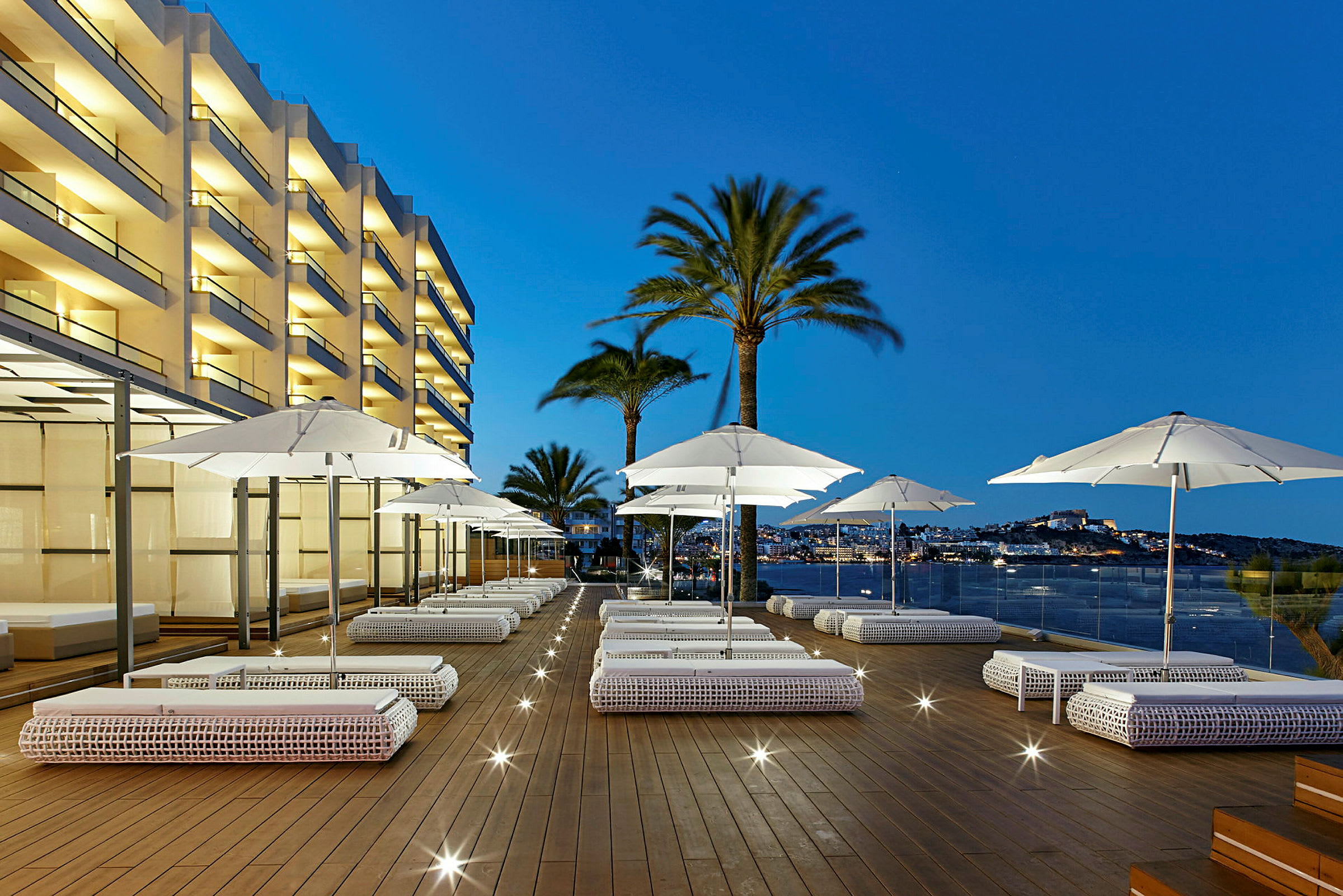 En hotellterrass på kvällen med utomhussoffor, vita parasoller och utsikt över hav och stad. (AI-genererad text)