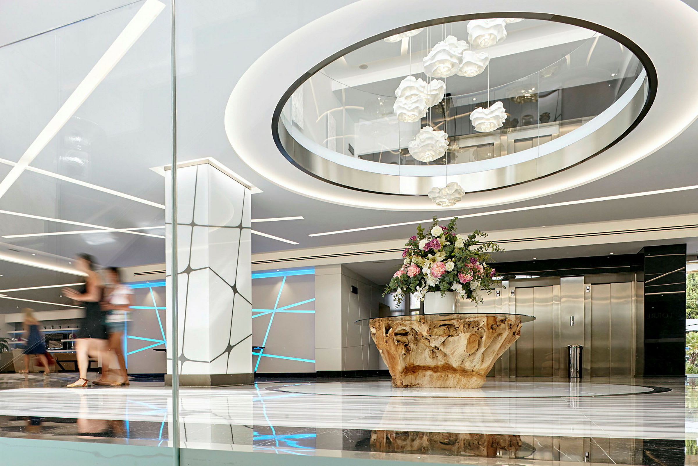 Ljus och modern hotellobby med ett stort bord av trä och en bukett blommor i mitten, eleganta ljuseffekter och hissar i bakgrunden. (AI-genererad text)