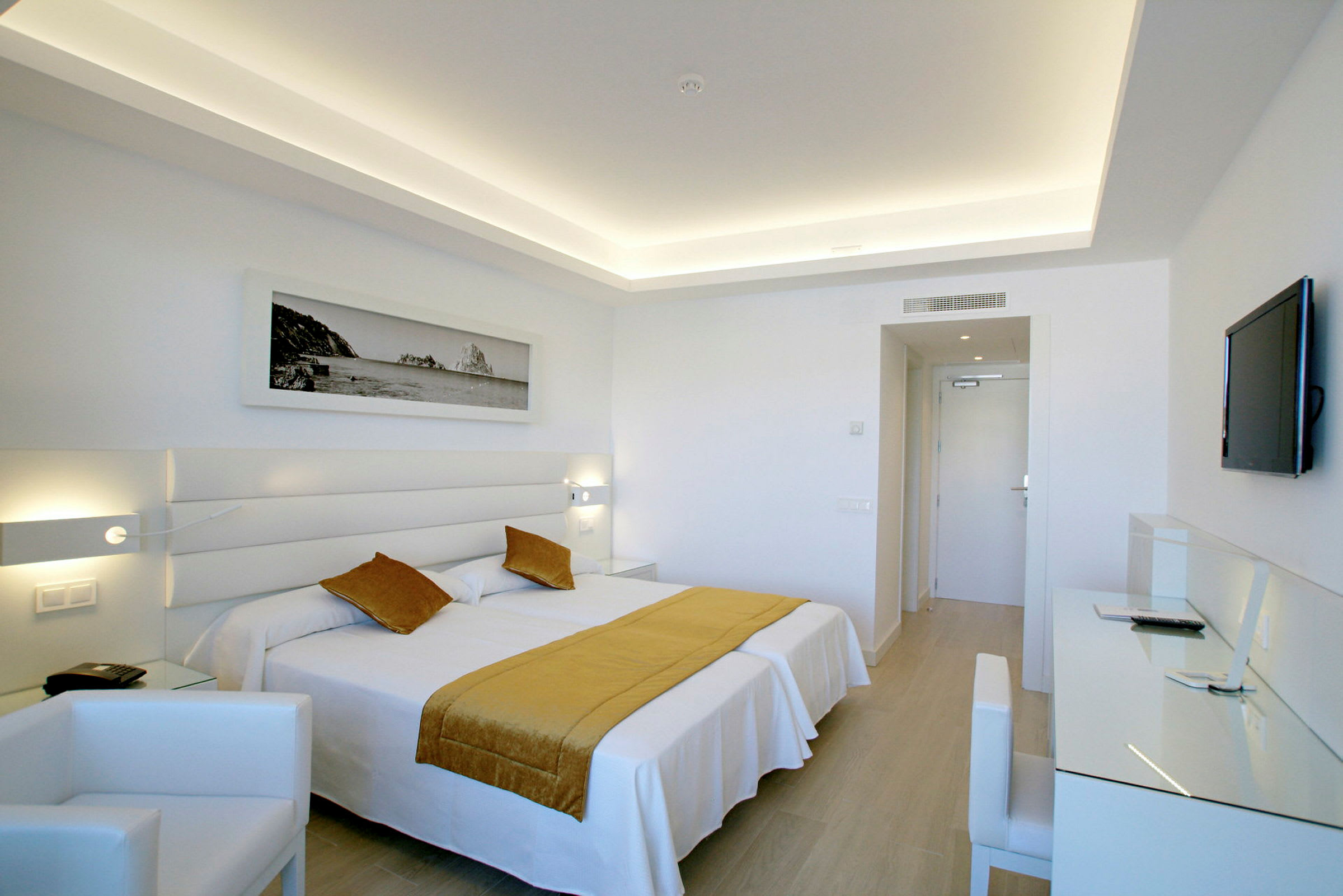 Ett modernt hotellrum med en dubbelsäng klädd i vit sängklädsel och rustika gula kuddrön och plädar. Rummet har en vit inredning med en skrivbordsplats, vägg-TV och konstverk ovanför sängen. (AI-genererad text)