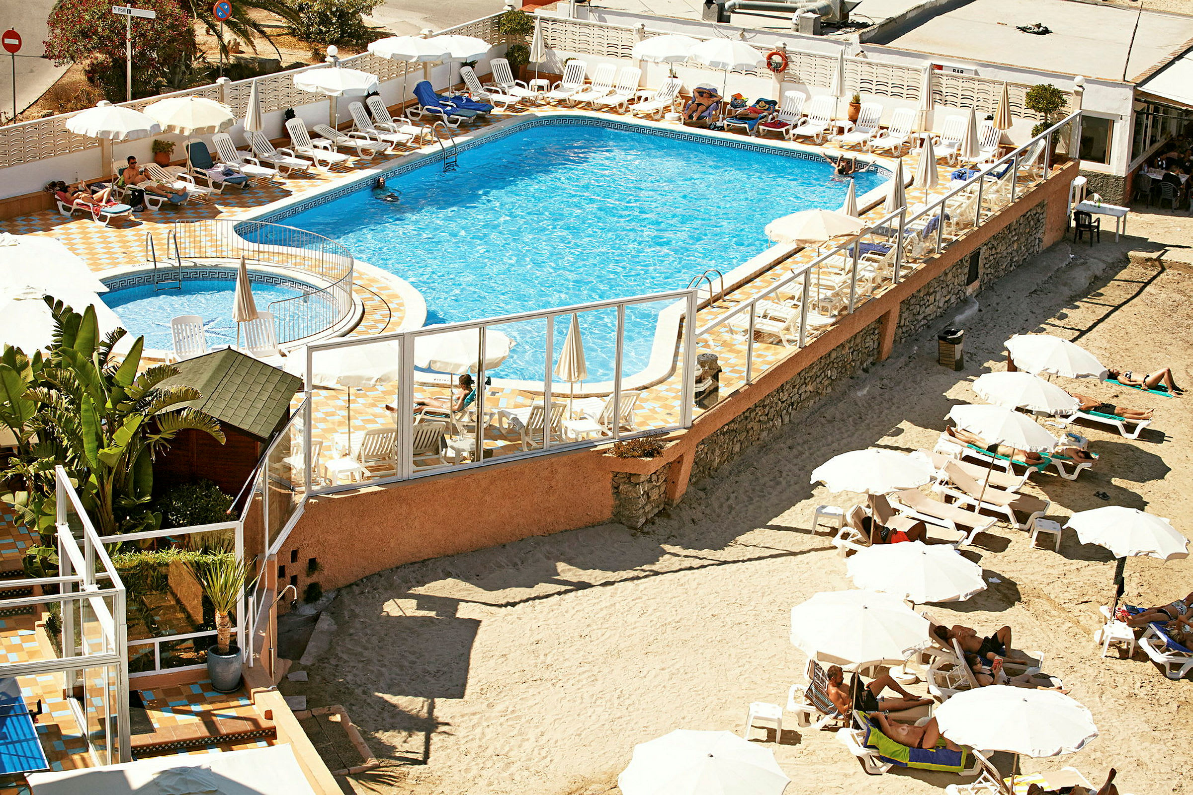 En pool omgiven av solstolar och parasoller, med en sandig yta och fler solstolar utanför området. (AI-genererad text)