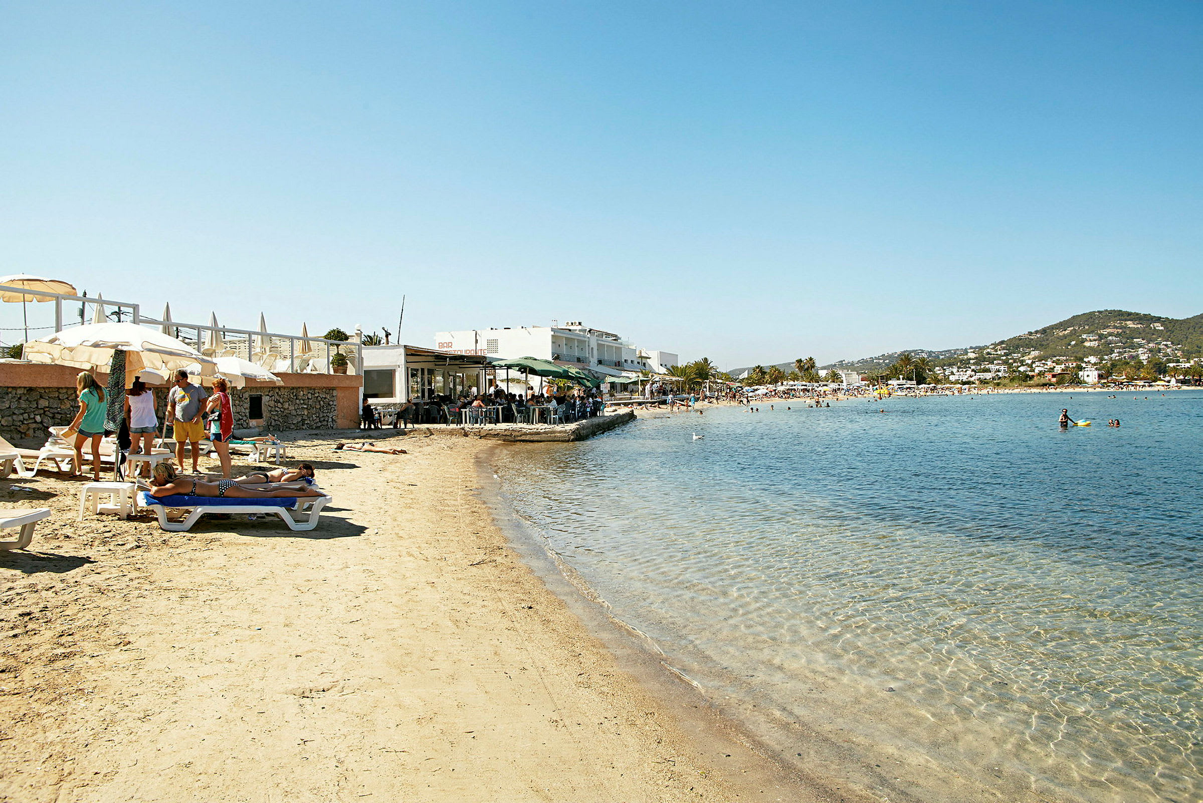 En sandstrand med människor som solar och badar. I bakgrunden finns en rad restauranger och byggnader. (AI-genererad text)