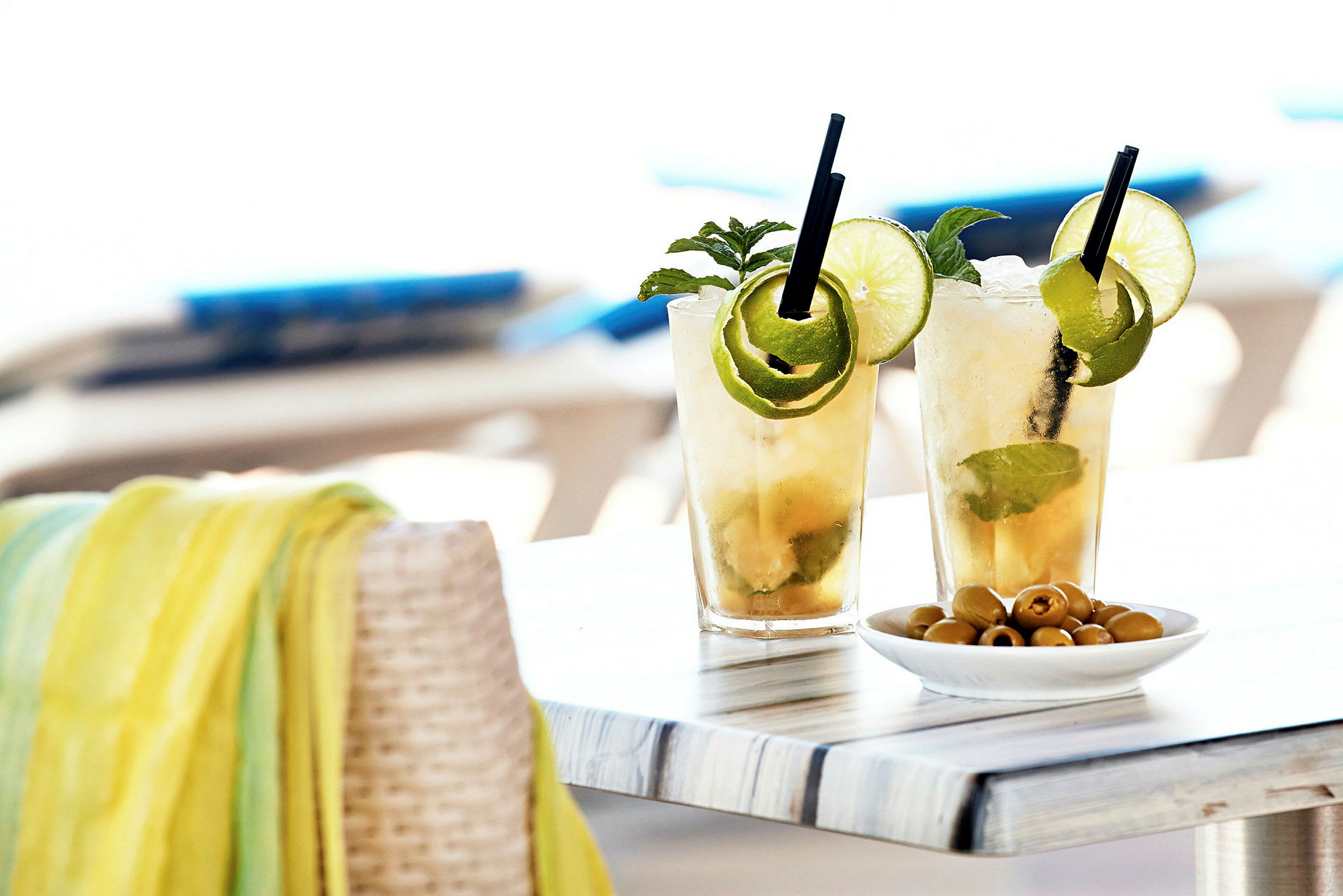 Två glas med kylda cocktails dekorerade med lime och mynta står på ett bord bredvid en skål med oliver. (AI-genererad text)