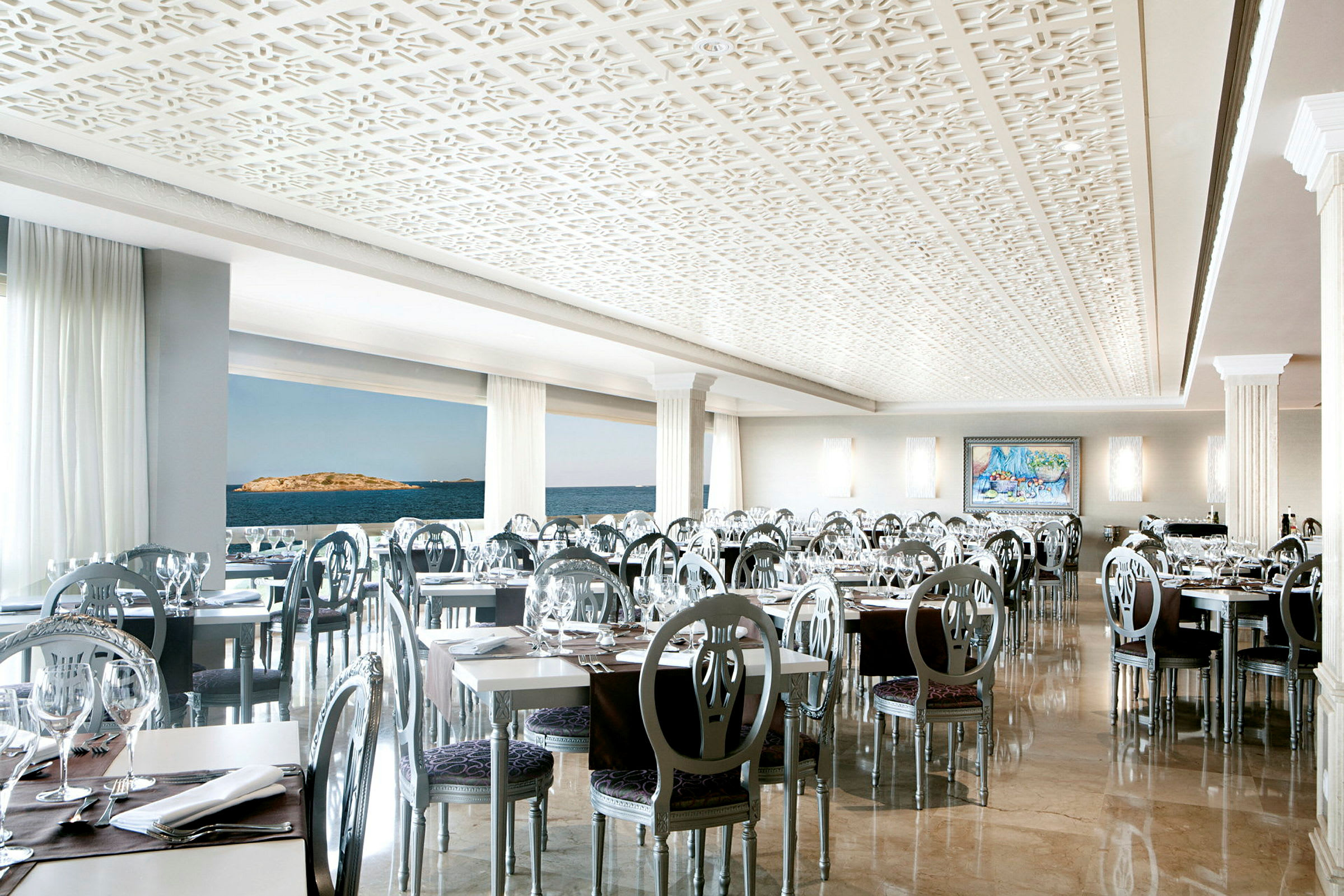 Ett elegant inrett restaurangutrymme med moderna stolar och vita bord, med stora fönster som visar en utsikt över havet. (AI-genererad text)