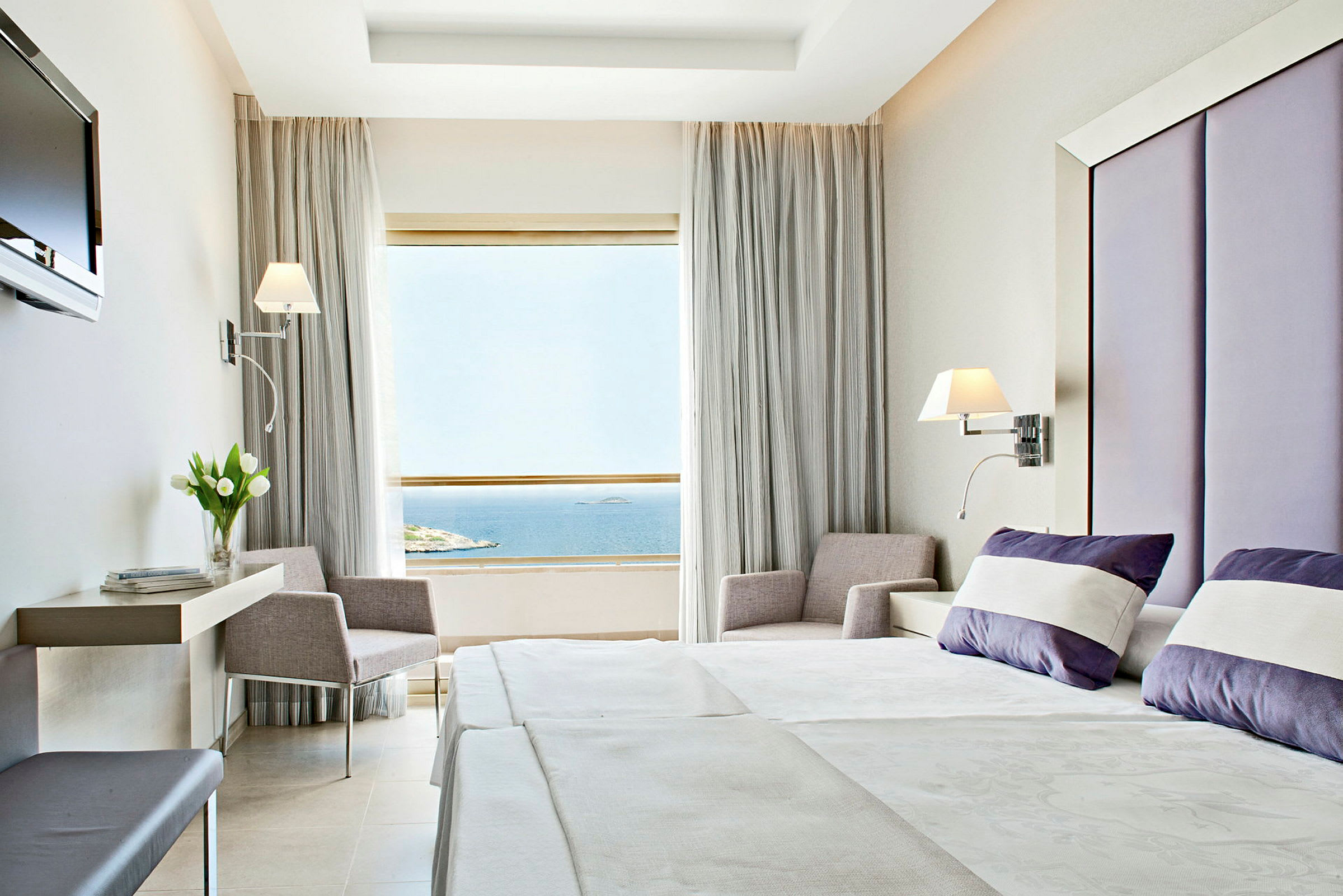 Ett hotellrum med modern inredning. Sängen är bäddad med vita och lila kuddar, och ett stort fönster med utsikt över havet visas. (AI-genererad text)