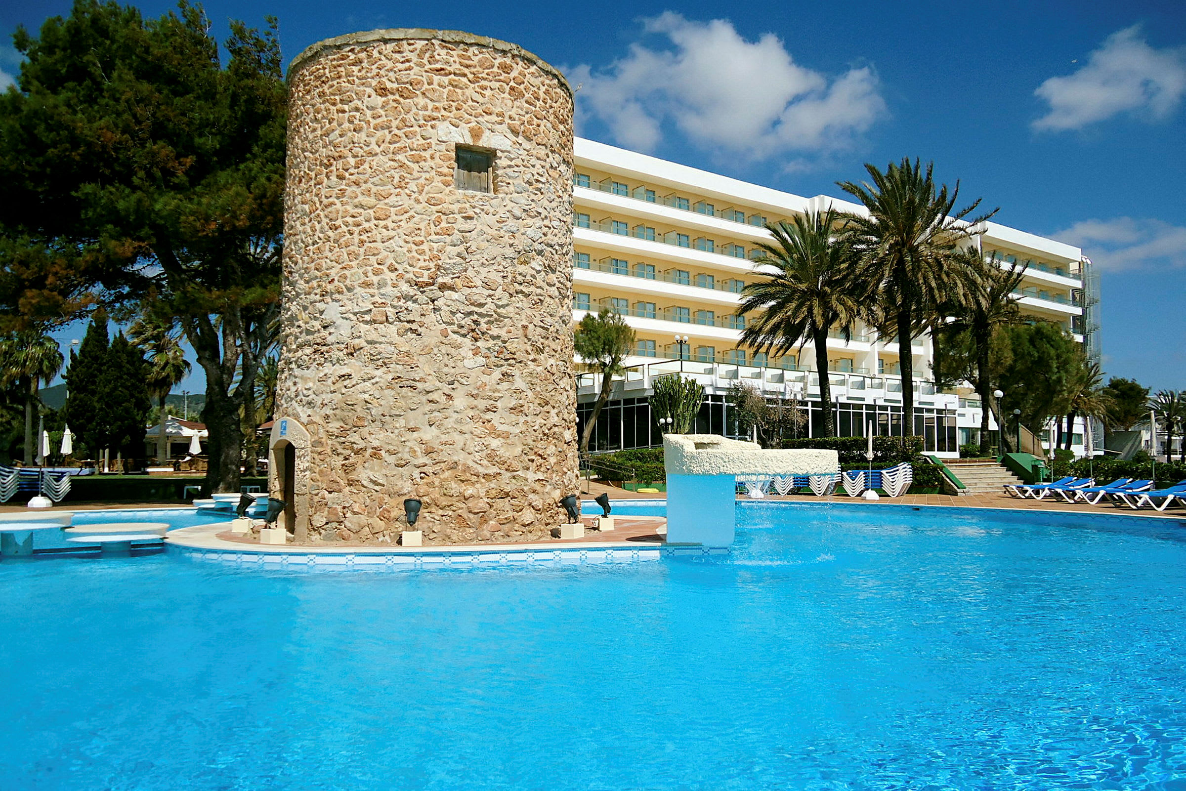 En stenbyggnad vid kanten av en klarblå pool med ett hotell i bakgrunden. (AI-genererad text)