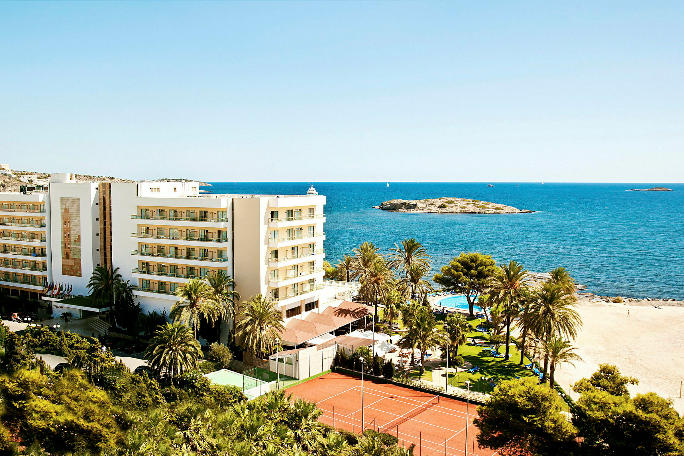 Bild av en semesteranläggning intill stranden med en byggnad, palmträd, pool och tennisbanor, belägen vid havet. (AI-genererad text)