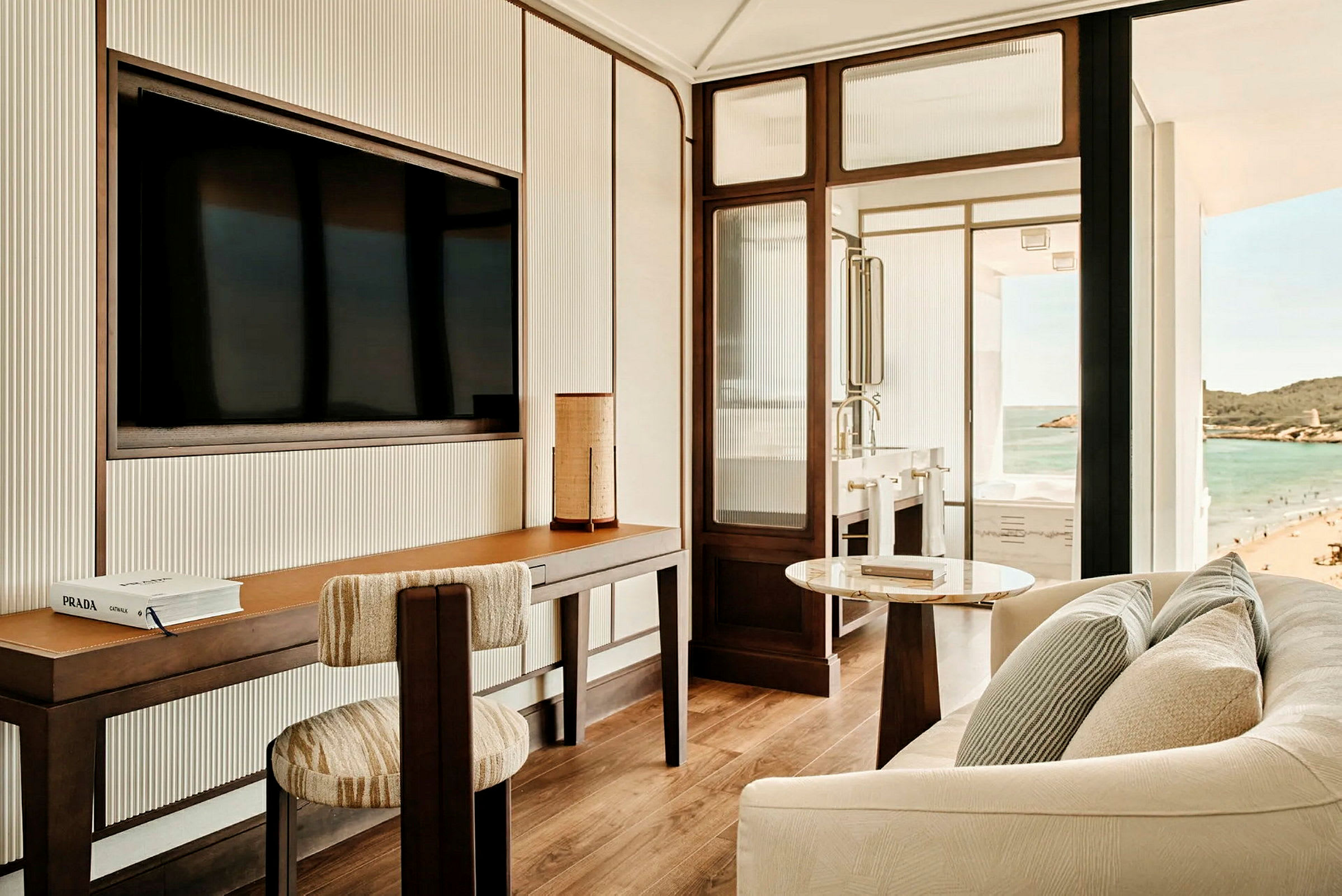 Ett modernt hotellrum med skrivbord, stol, soffa och utsikt över stranden och havet från fönstret. (AI-genererad text)