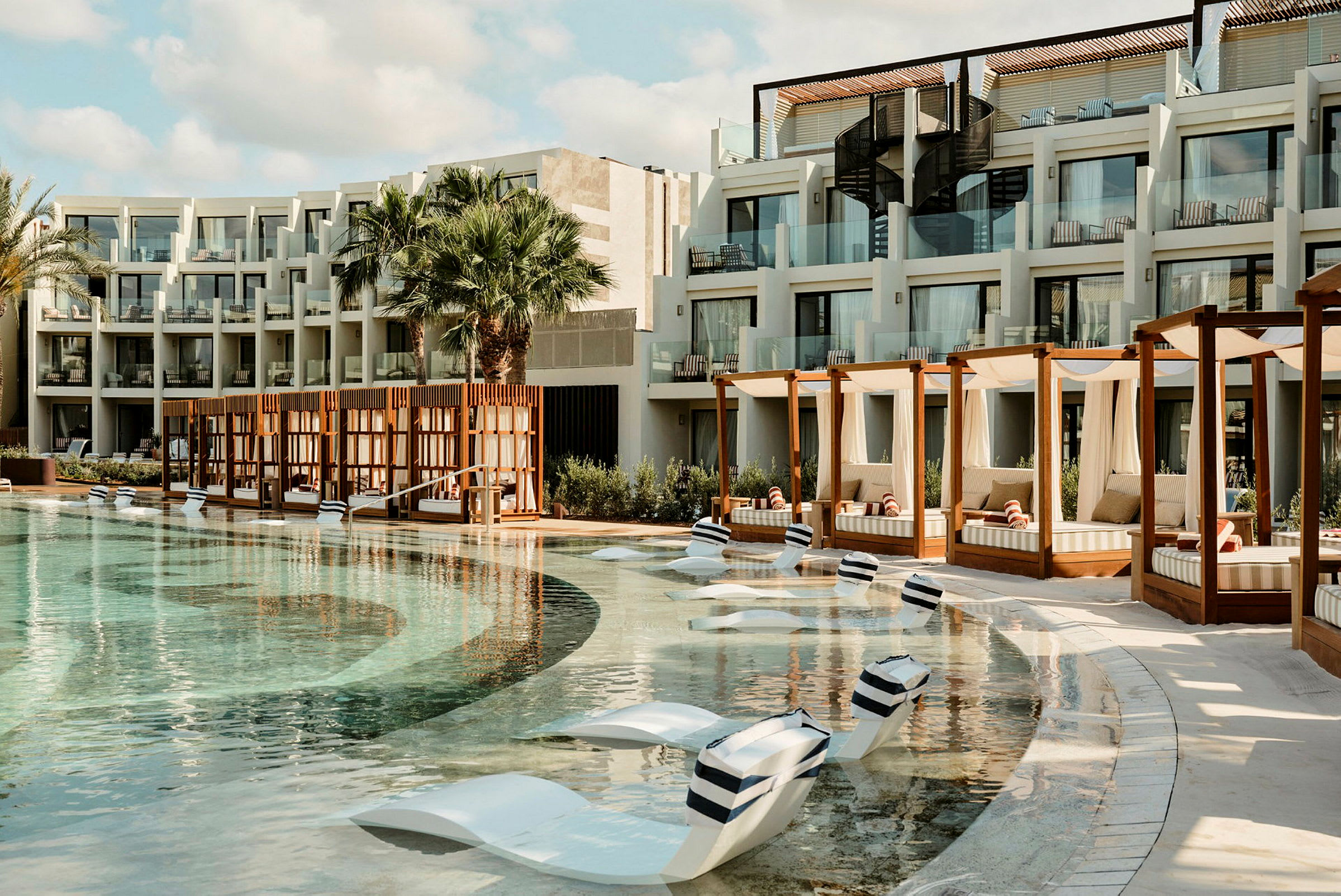 En hotellpool med solstolar i poolen och cabanas på sidan. Byggnaden har balkonger med glasräcken. (AI-genererad text)