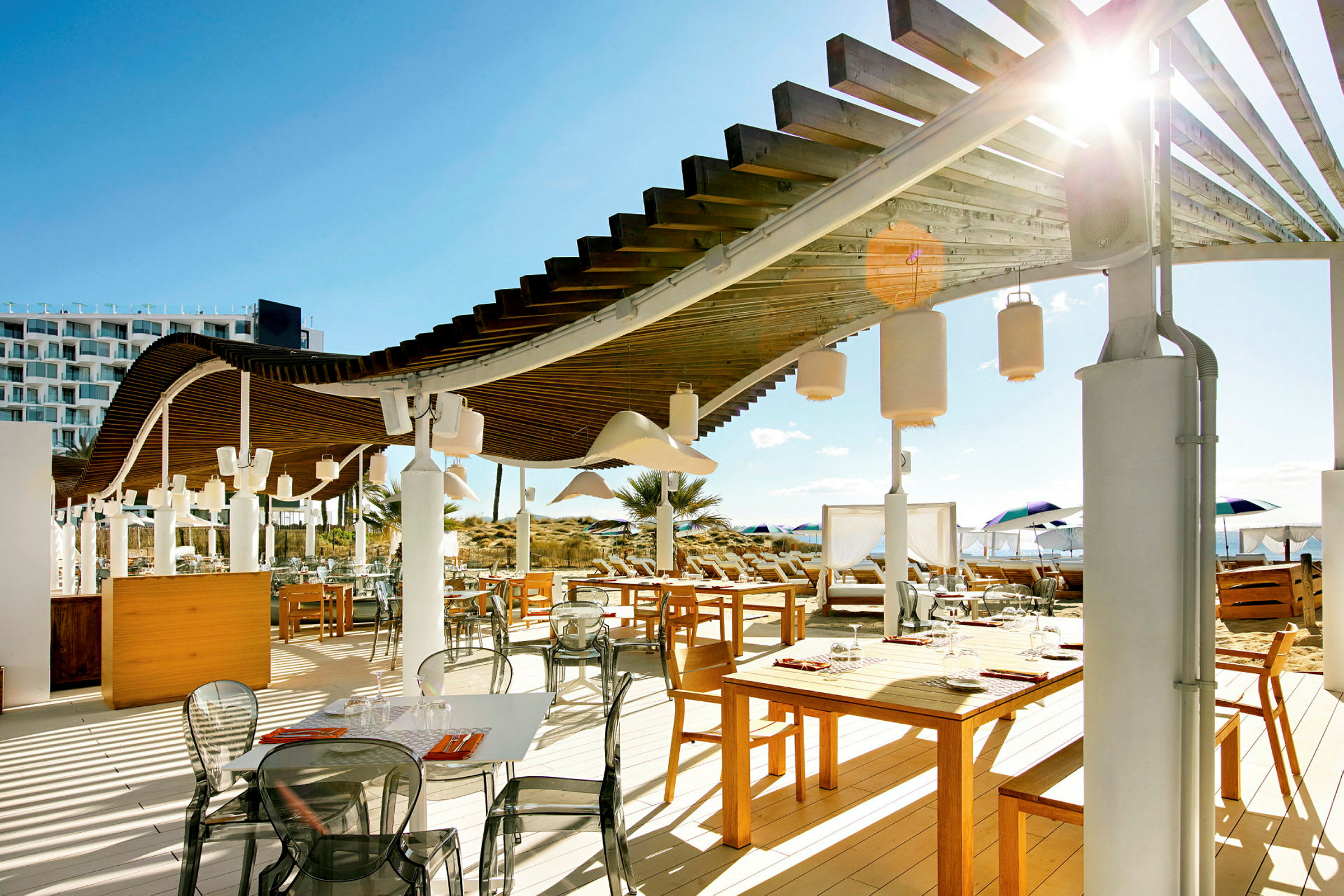 En utomhusrestaurang med träbord, stolar och en pergola över. I bakgrunden syns strand och parasoller. (AI-genererad text)