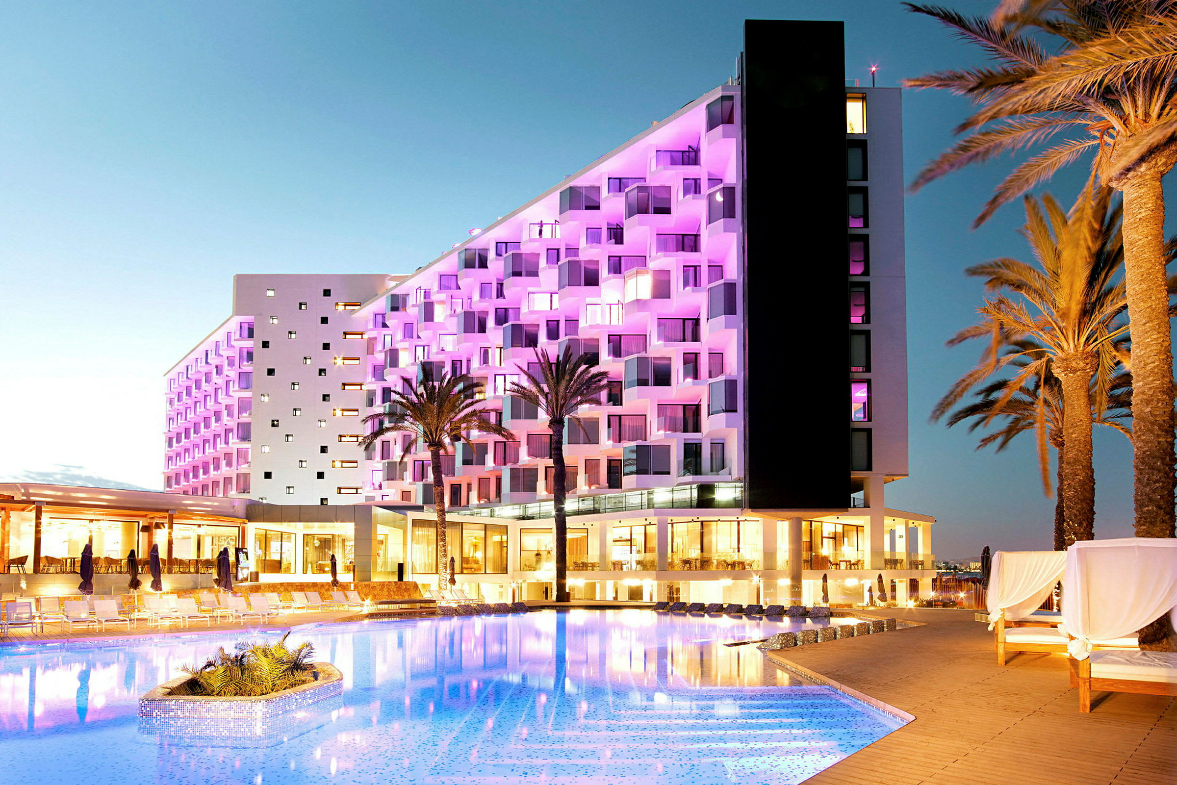 Ett hotell med fasad belyst med rosa ljus intill en pool, omgiven av palmträd. (AI-genererad text)