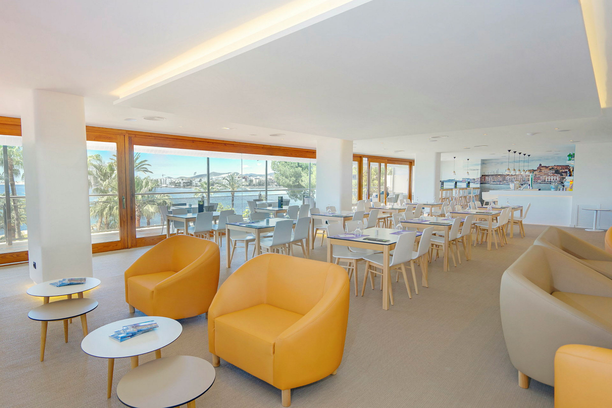 Ett ljust café eller restaurangområde med stora fönster som släpper in naturligt ljus, sittgrupper i moderna stolar och bord. (AI-genererad text)