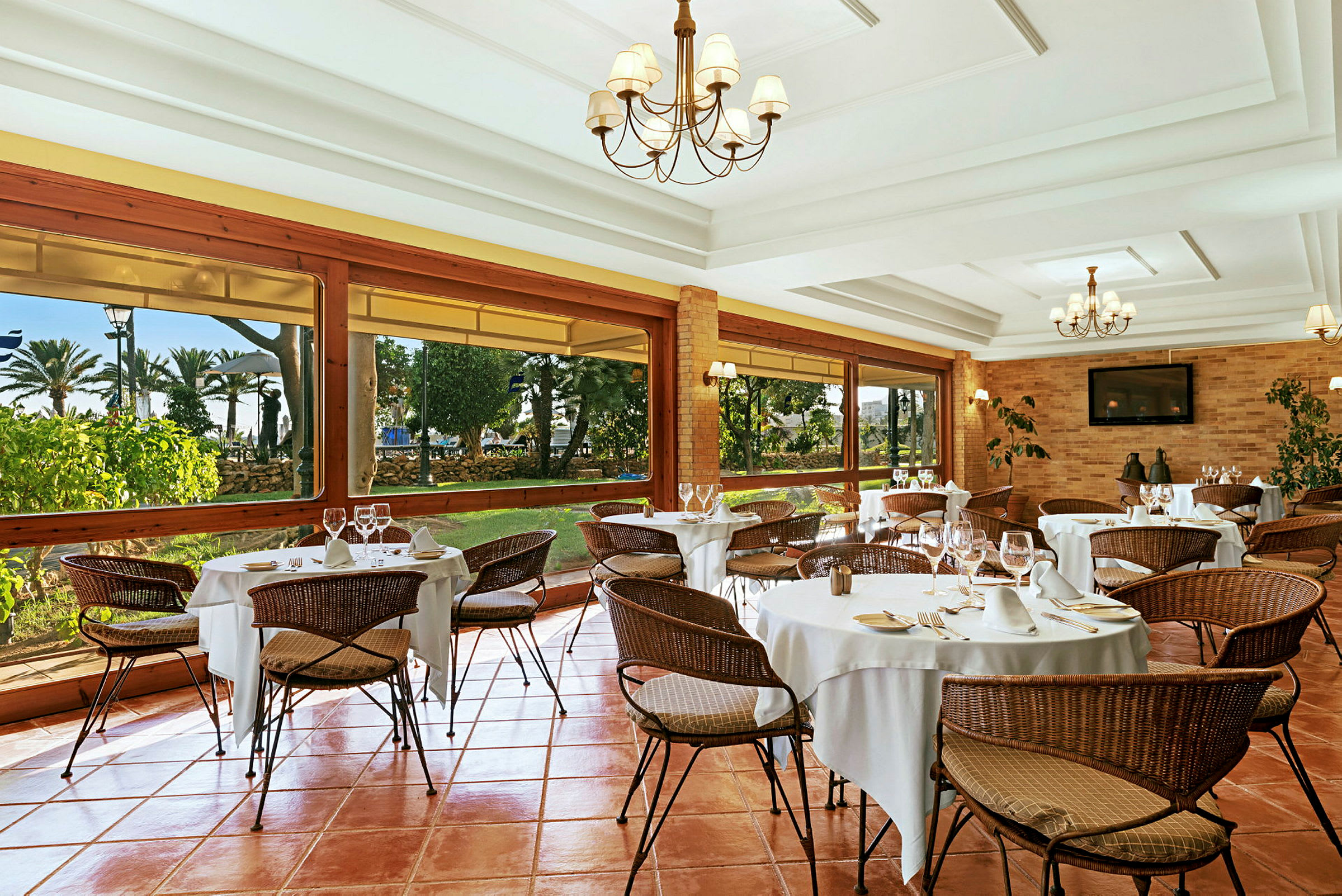En elegant restaurang med dukade bord och flätade stolar. Stora fönster visar en utsikt över en trädgård. (AI-genererad text)