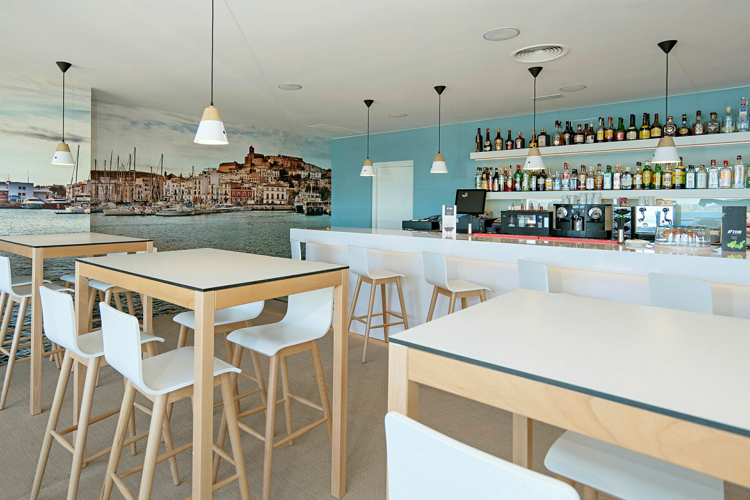 Ett modernt kafé med höga bord och stolar, en bar med drycker i bakgrunden och en stor väggmålning av en hamnstad. (AI-genererad text)