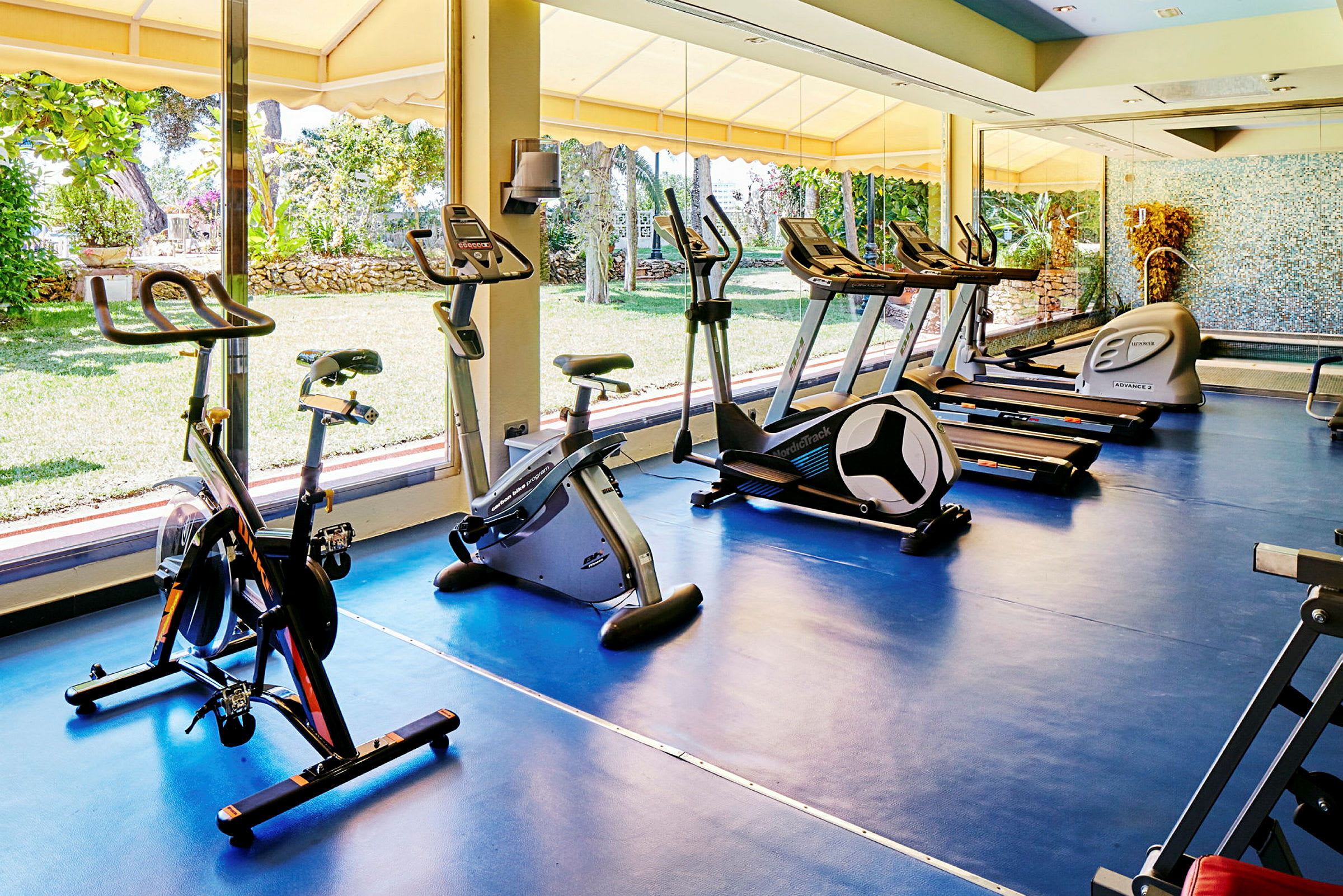 Ett gym med flera träningsmaskiner inklusive motionscyklar och löpband, placerade vid ett stort fönster med vy ut över en trädgård. (AI-genererad text)