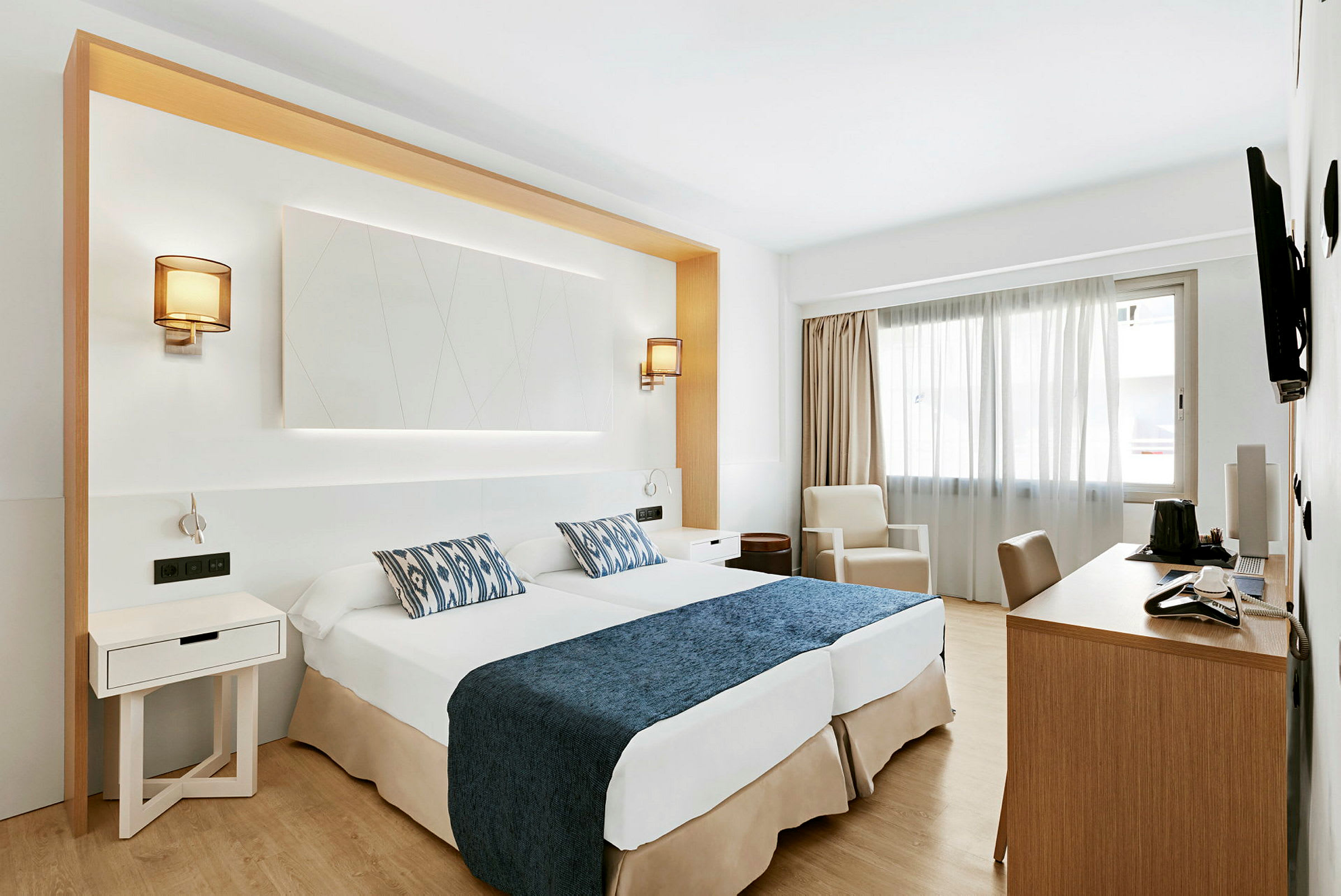 Ett hotellrum med två sängar försedda med blå överkast och mönstrade kuddar, skrivbord, stol och en fåtölj bredvid ett fönster. (AI-genererad text)